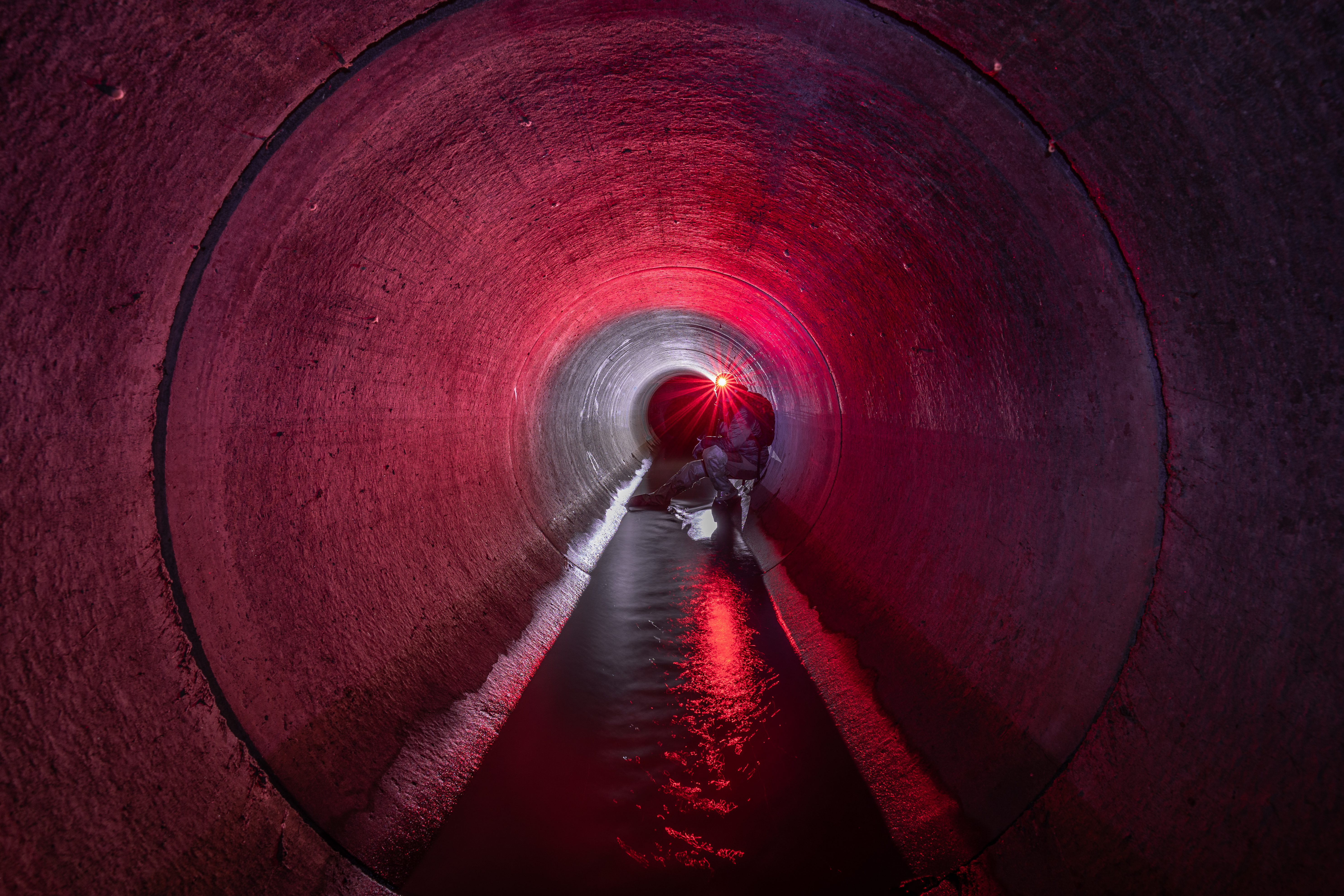 A dimly lit tunnel with a red glow, showing a crouching figure in the distance, reflecting light on water at the base.