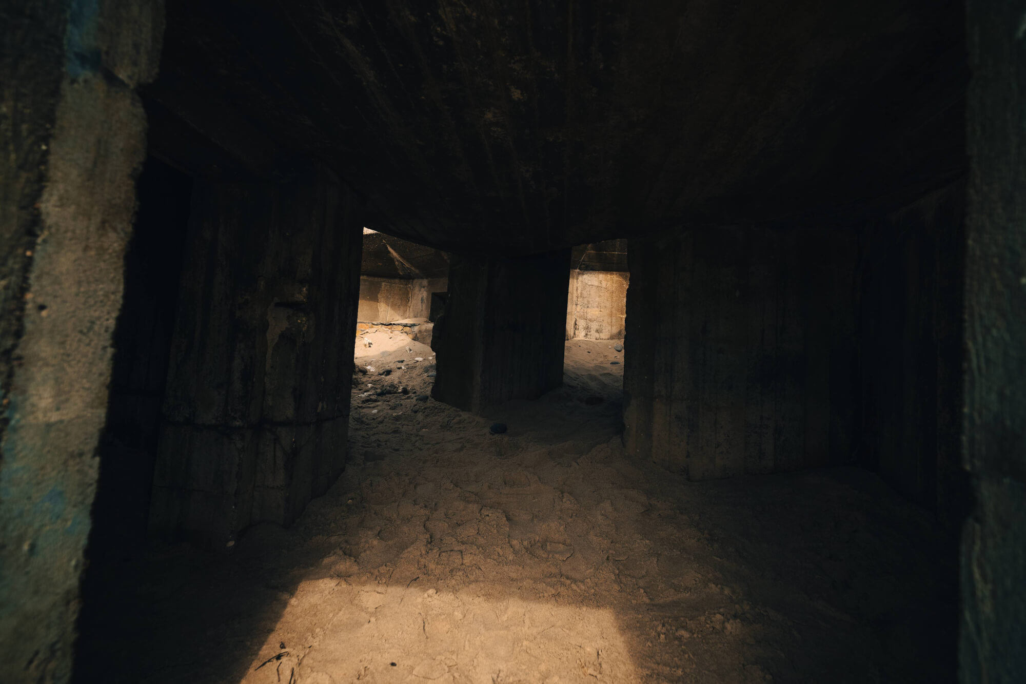 A dimly lit interior of a concrete structure with large supporting pillars, sandy ground, and shadows cast by the overhead elements.