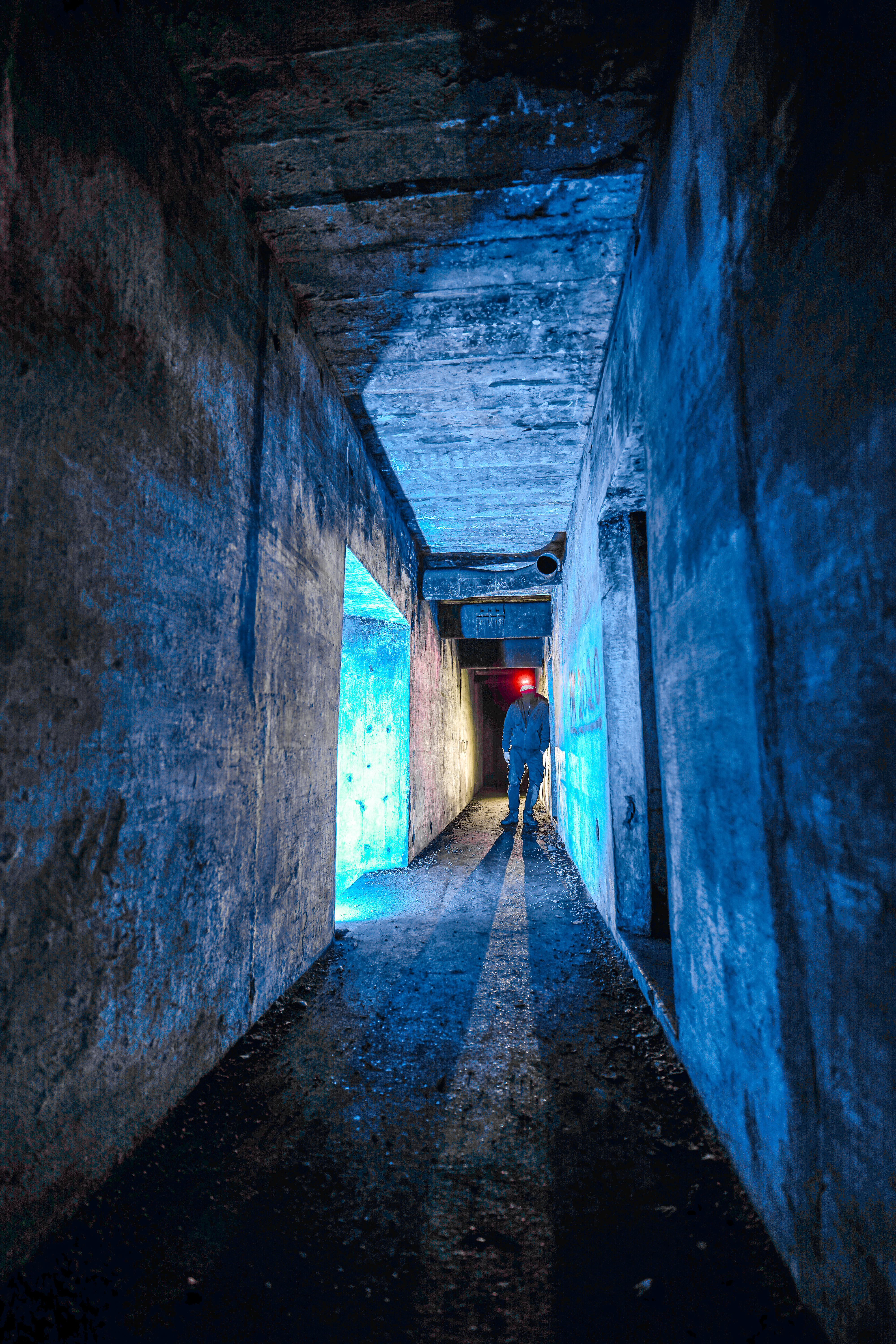 A dimly lit concrete corridor with a person standing at the end, illuminated by blue light and casting long shadows across the floor.