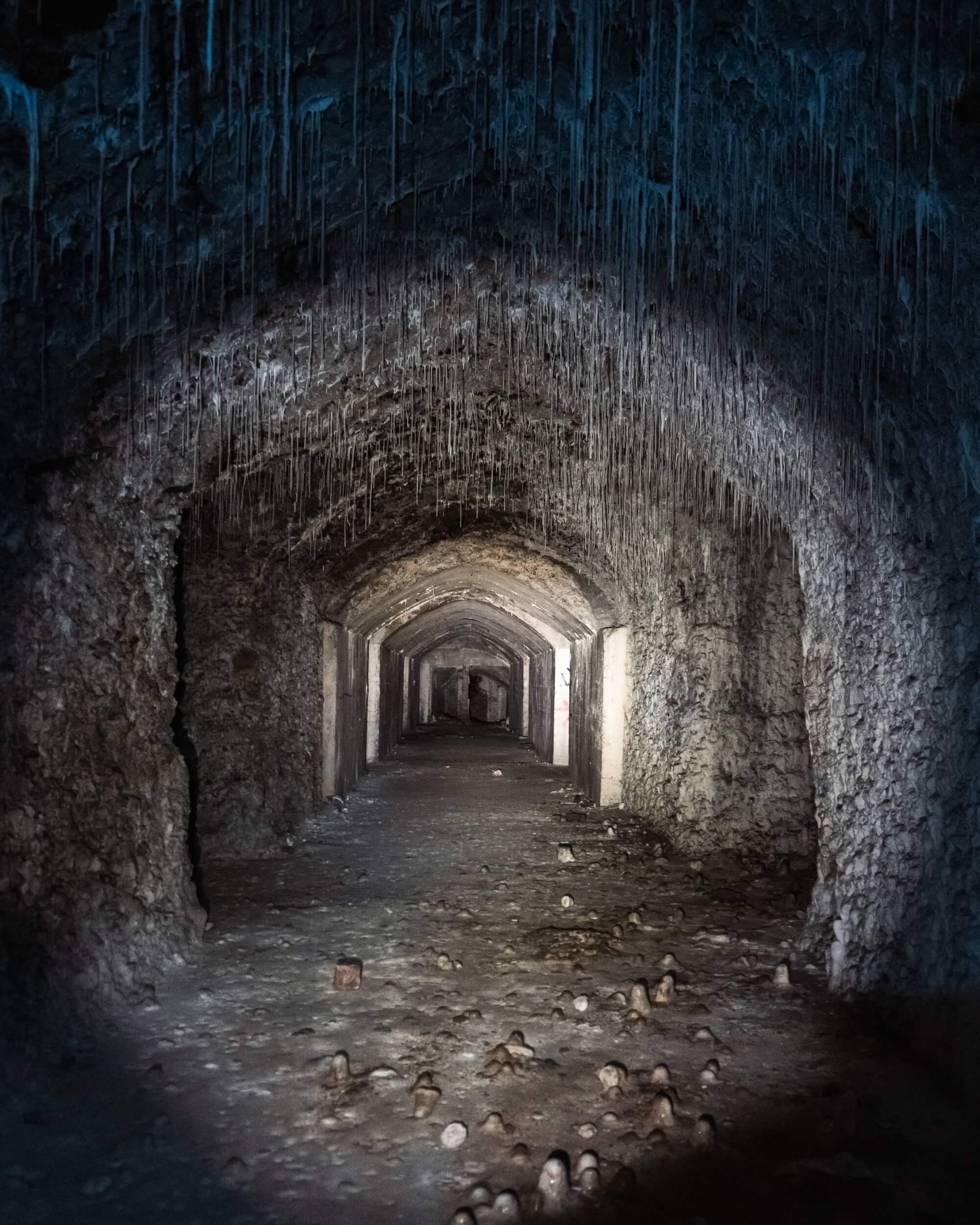 A dark, old stone corridor with stalactites hanging from the ceiling. The ground is uneven and littered with stones. The light illuminates part of the passage, creating a mysterious mood.