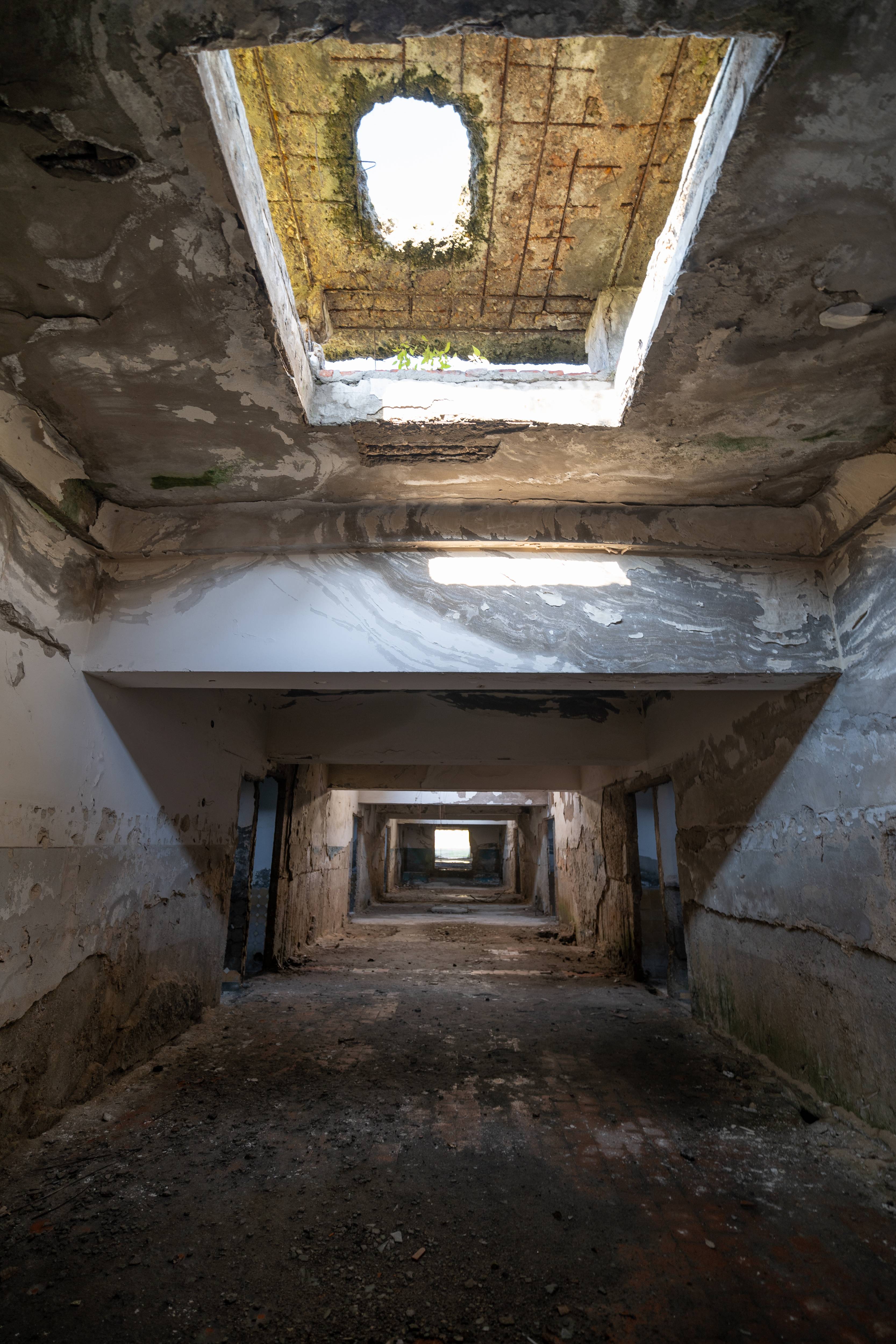 View of an abandoned corridor with peeling walls and a hole in the ceiling letting in light, exposing dusty ground and empty rooms in the foreground.