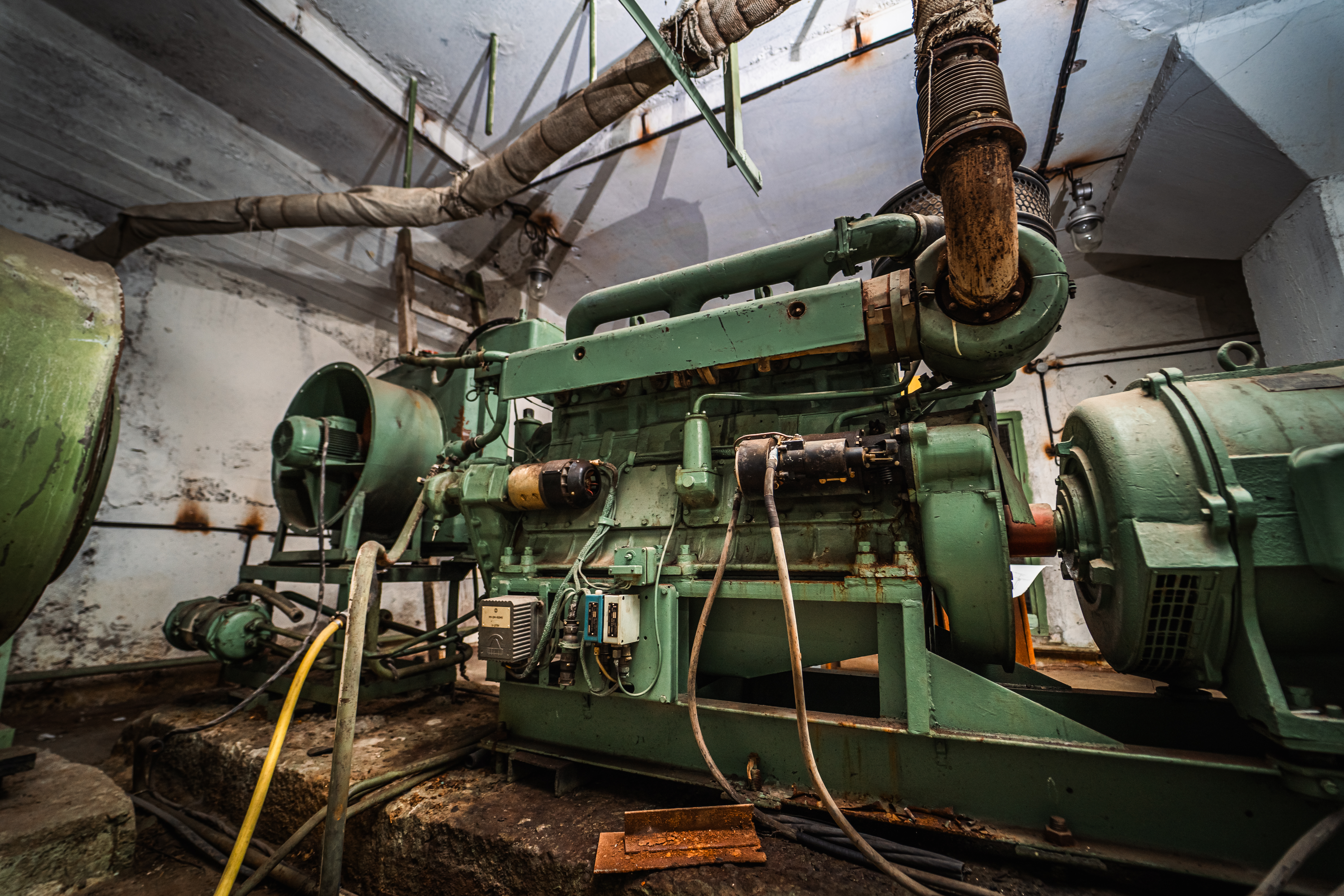 A large, green industrial generator with pipes overhead and rusted elements in a dimly lit room, suggesting abandonment and neglect.