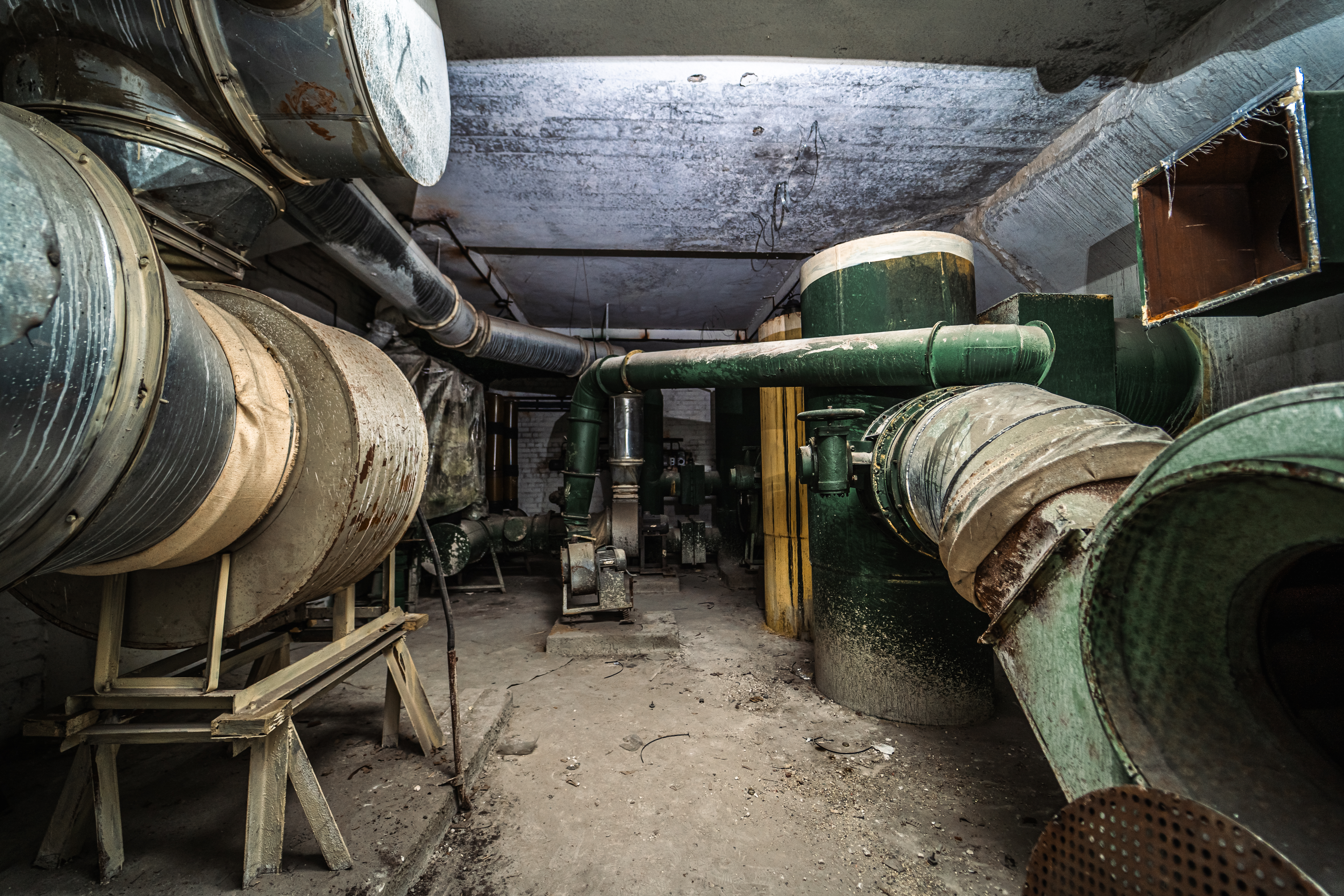 An abandoned industrial space with rusted green pipes and machinery, illuminated by soft overhead light, creating an atmosphere of neglect.