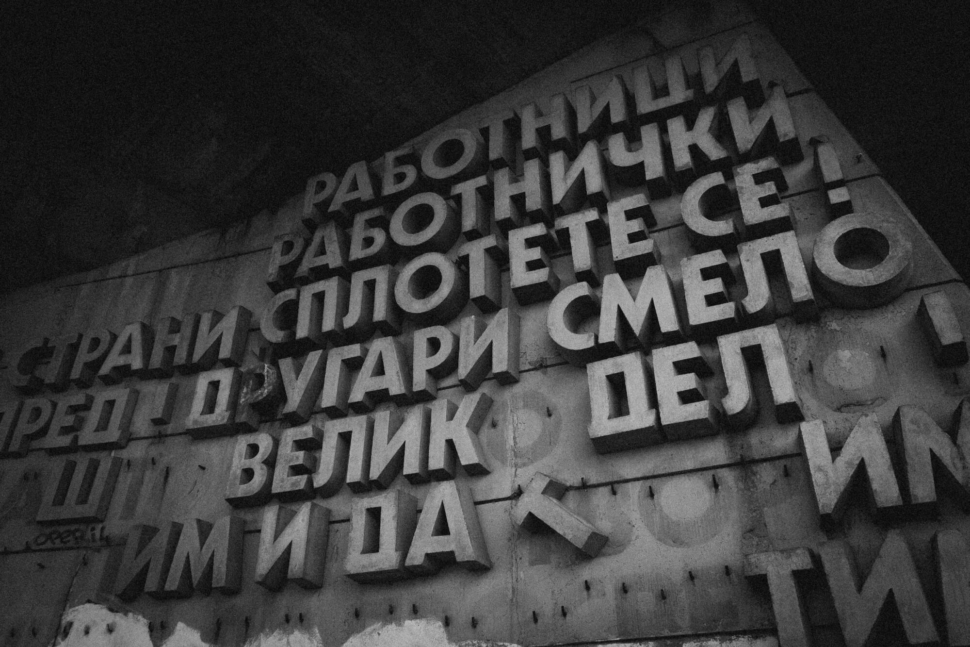 Close-up of a textured concrete wall with raised Cyrillic letters, conveying a message related to labor and solidarity. The image is in black and white, highlighting the contrast between the letters and the background.