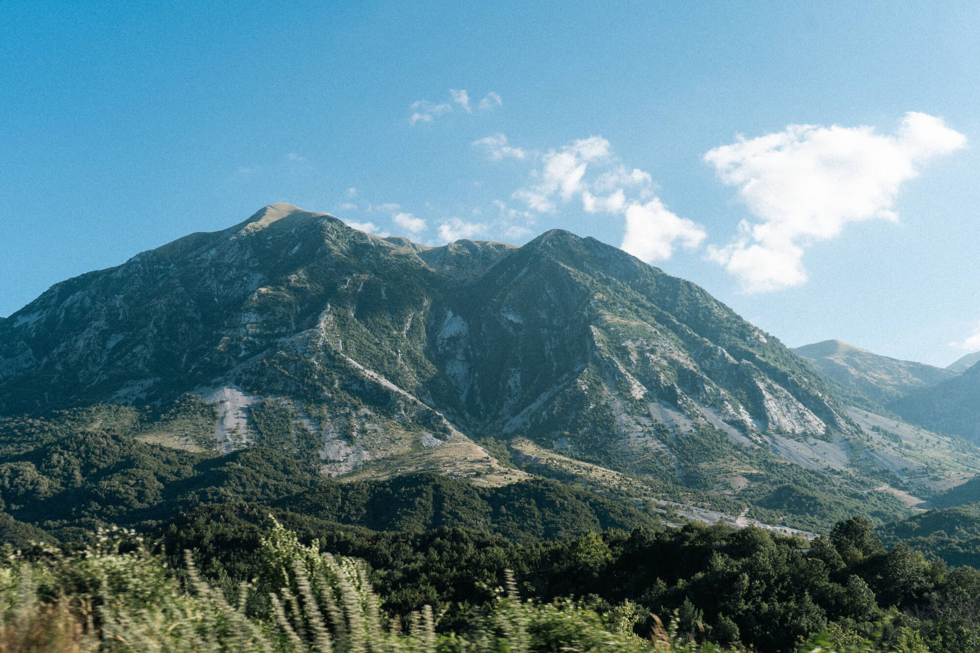 A towering mountain with rocky slopes covered in green vegetation under a clear blue sky with some clouds.
