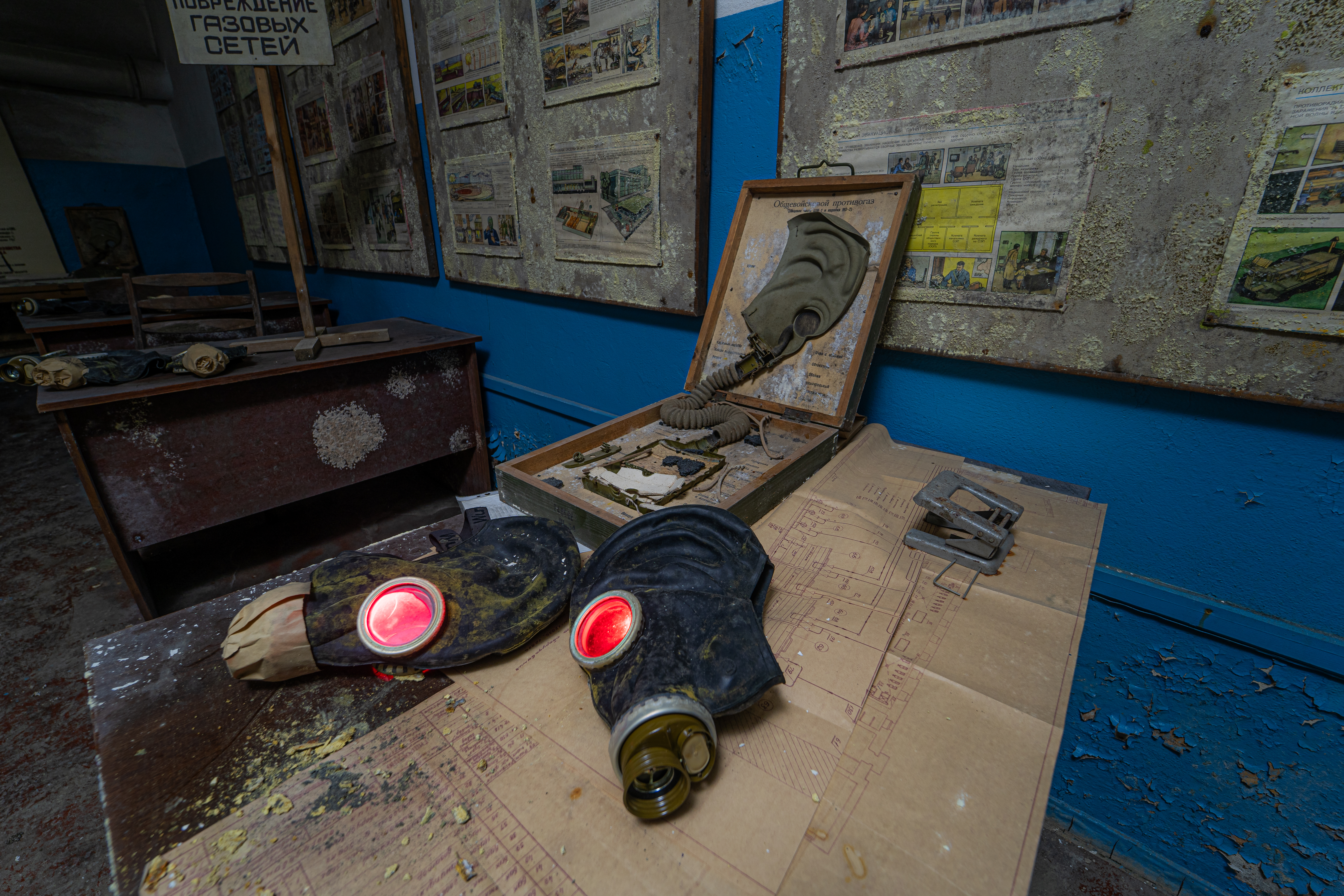 A close-up view of a vintage gas mask with red lenses resting on a desk, alongside an opened box containing another gas mask. The background features walls with faded posters and a wooden desk in low light.