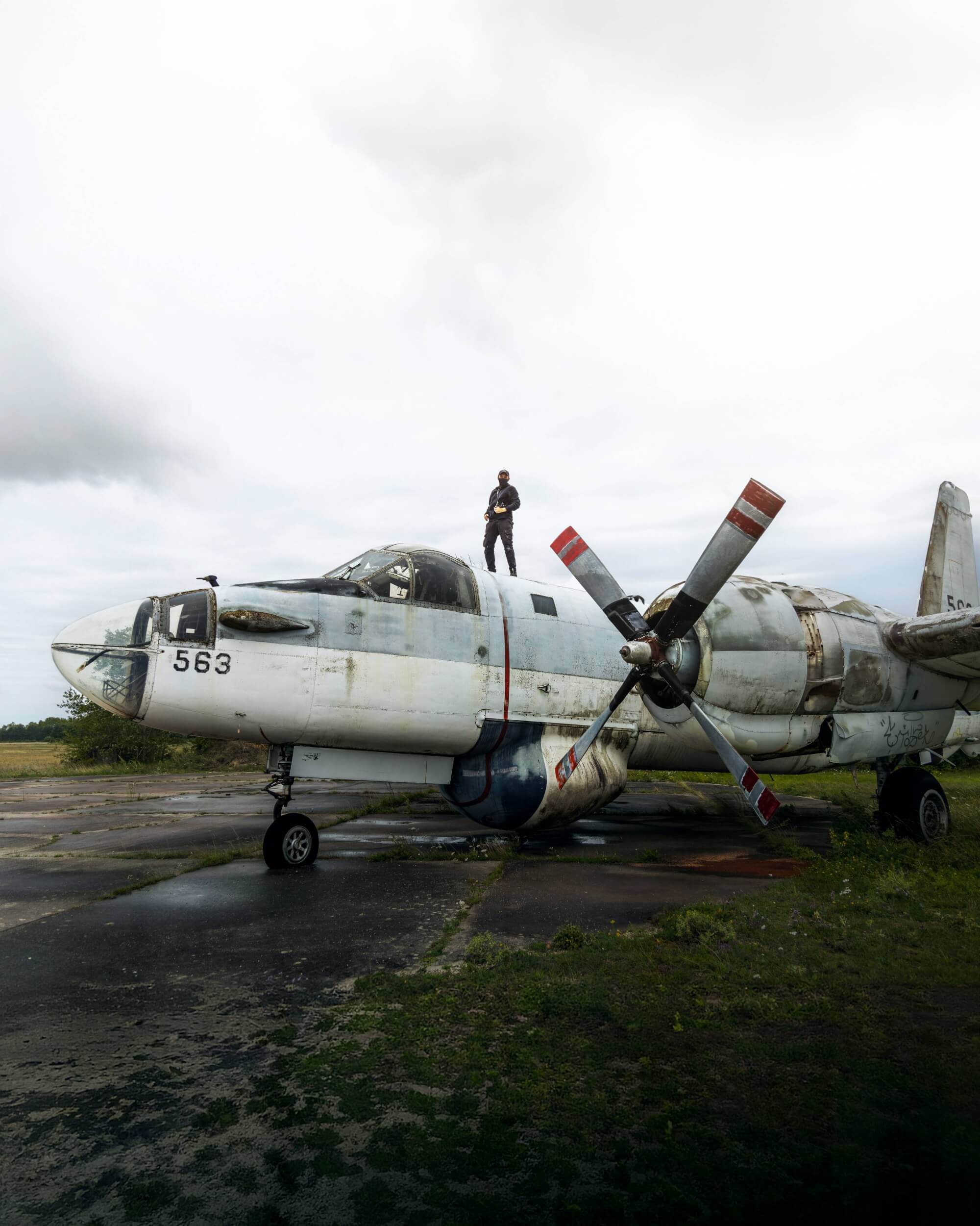 A worn-out airplane with a man standing on top, against a cloudy sky and surrounded by an overgrown airstrip.