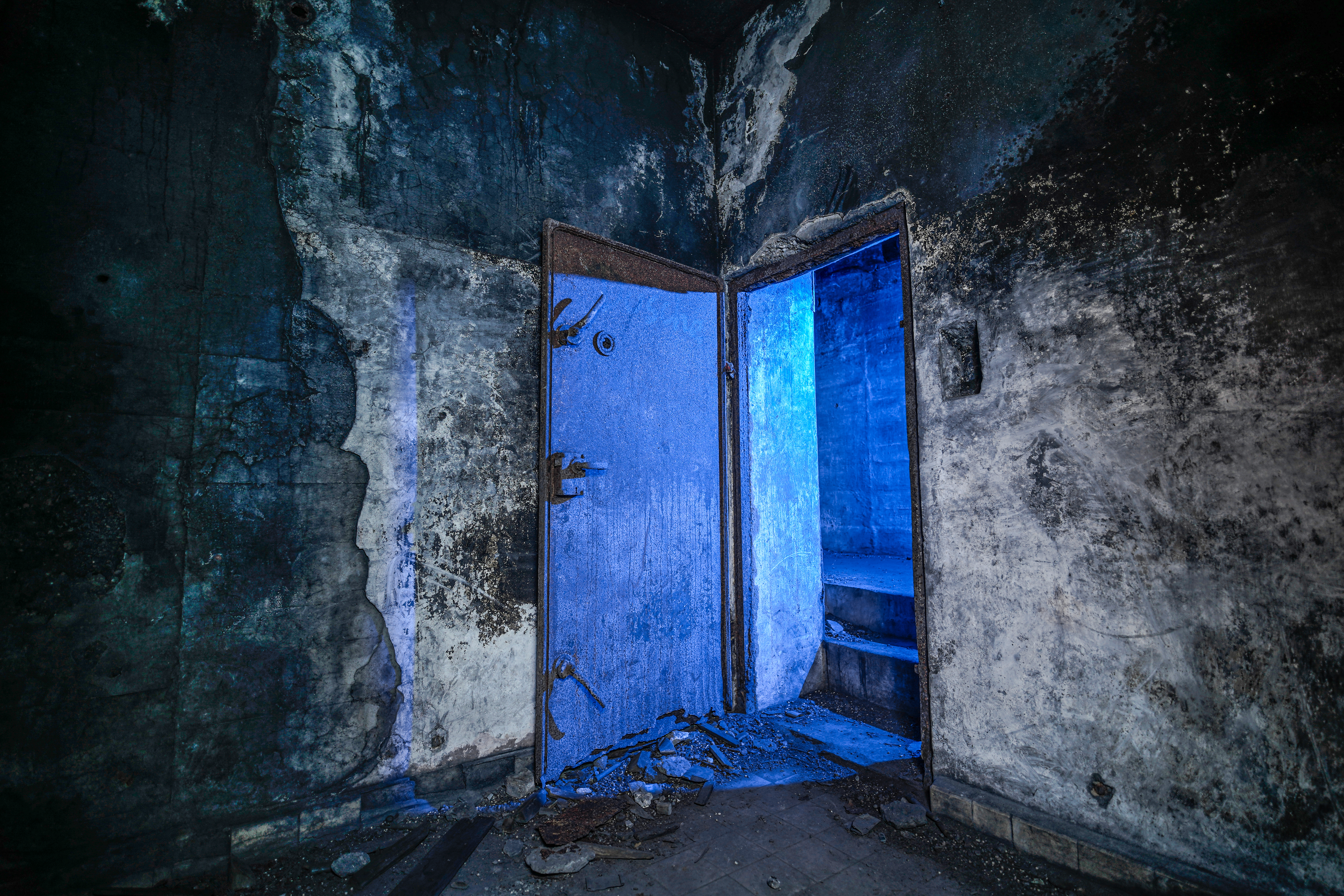 A partially open rusted iron door in a dark, distressed room, illuminated by a blue light. The walls are covered in soot and grime, with debris on the floor.