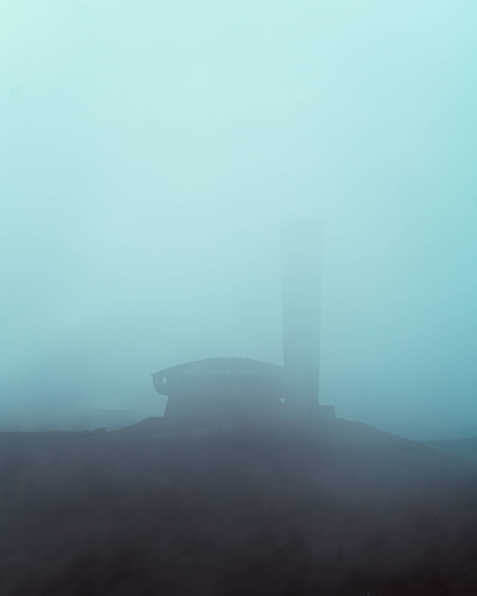 A foggy landscape with a dark silhouette of a building and a tall chimney emerging from the mist.