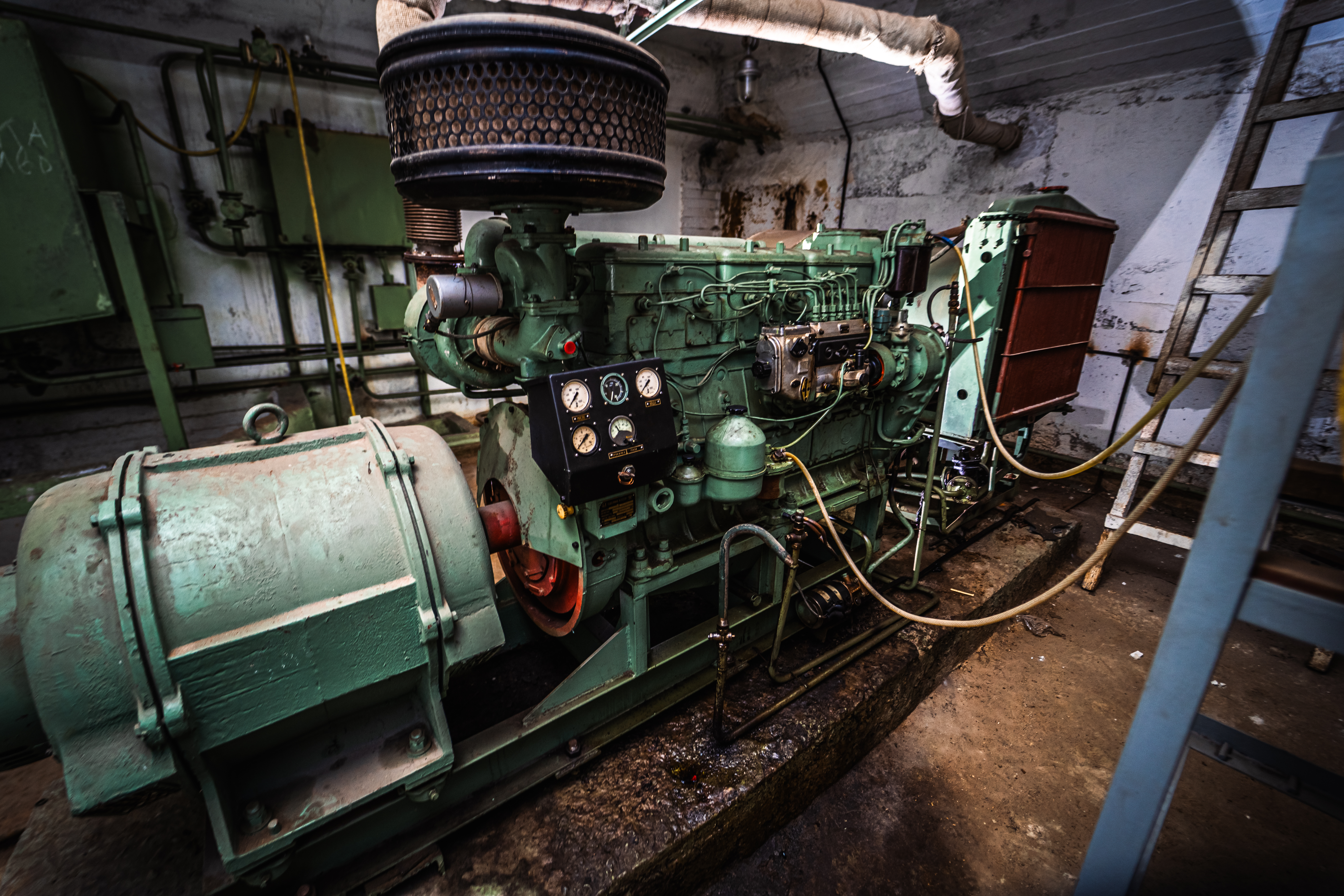 An industrial engine with various gauges and piping, set in a dim room with peeling walls and a wooden ladder in the background.