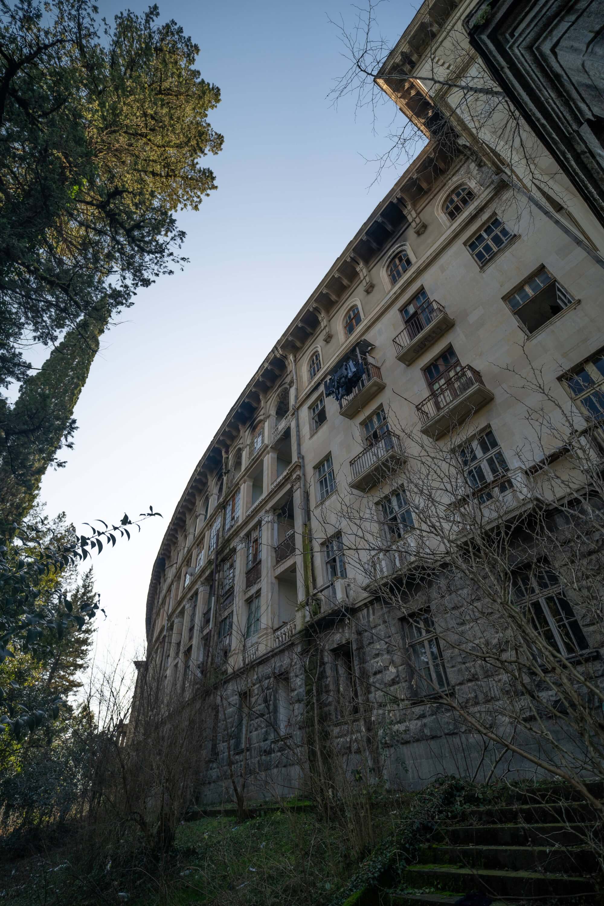 A tall, abandoned curvilinear building surrounded by trees and overgrown vegetation, viewed from a low angle. It has balconies and windows, some of which are broken. The sky above is clear and blue, while remnants of clothing hang on a balcony. The foreground features tangled, dry branches and grass.