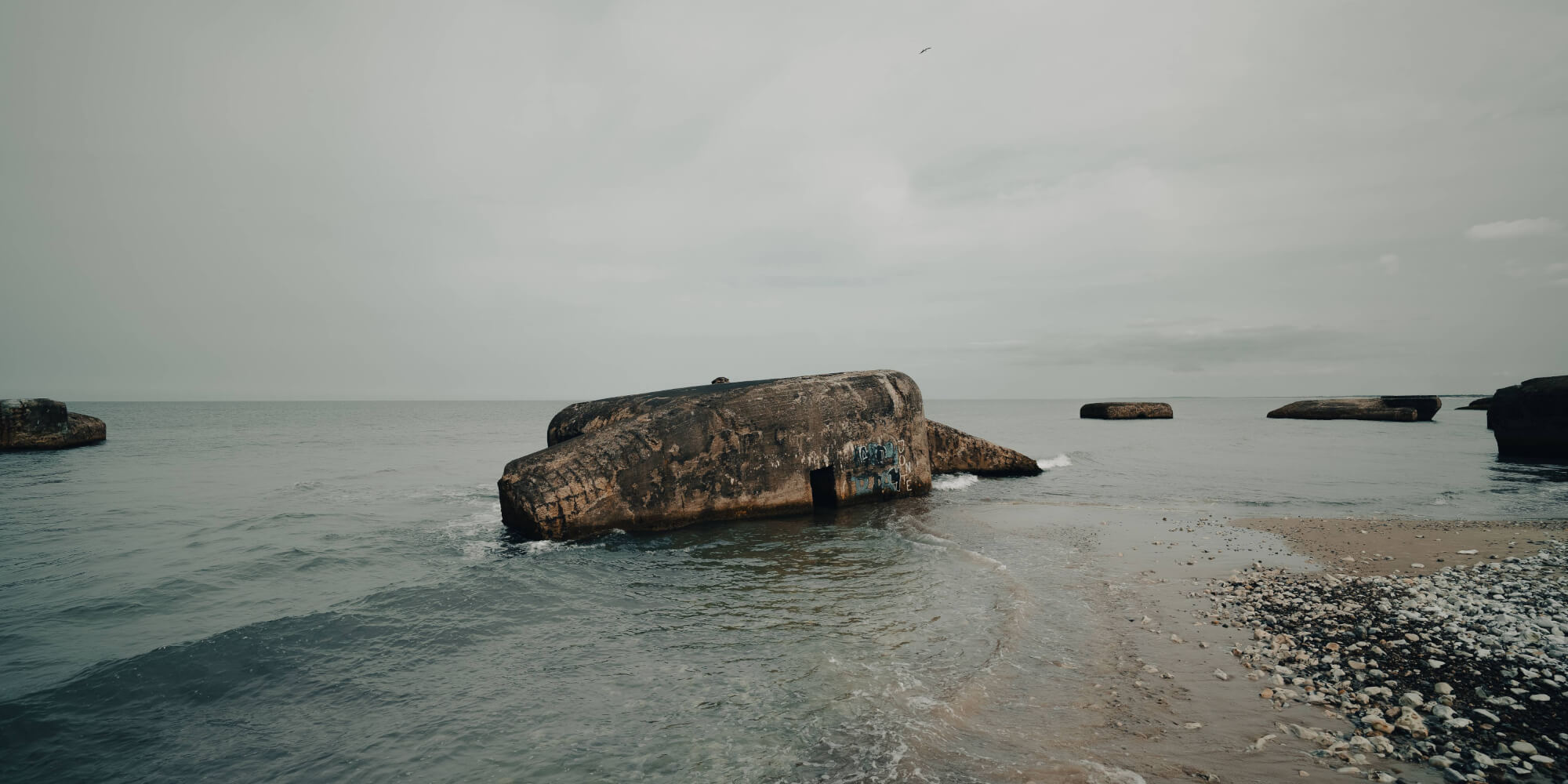 A coastal scene featuring large, weathered rock formations partially submerged in calm waters, with a smooth sandy shore and scattered pebbles. The sky is overcast, creating a soft, gray ambiance.