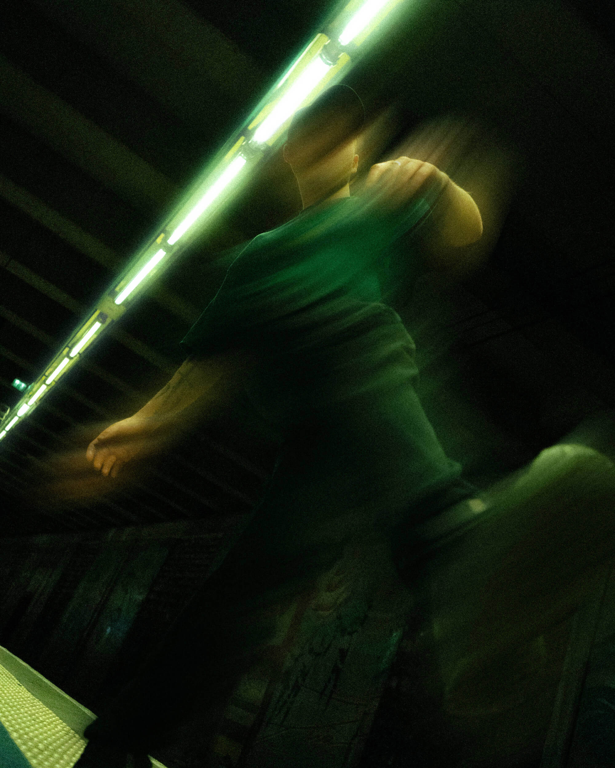 A blurred figure in a green shirt is captured mid-stride, under fluorescent green lighting in an urban setting. The background is dark, enhancing the motion and energy of the scene.
