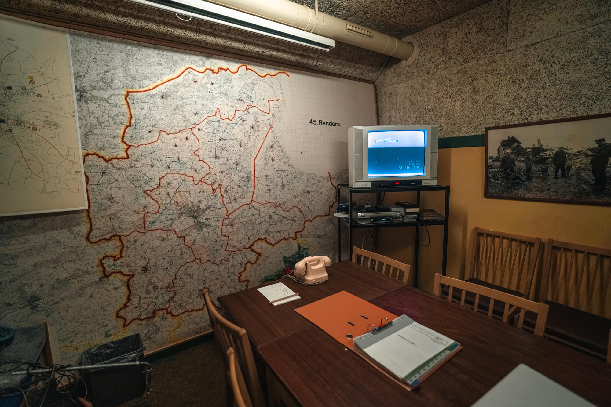 Interior of a strategy room featuring a large red-outlined map on the wall, a wooden table with documents and a pink telephone, a vintage television, and a framed photograph of people near wreckage.