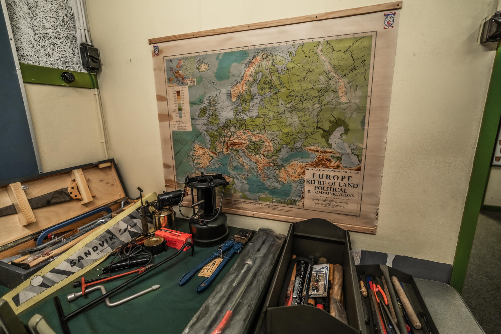 A workspace featuring a variety of tools on a green surface, with a detailed map of Europe pinned to the wall above. The tools include a saw, pliers, a lantern, and various other instruments, suggesting a functional and creative environment.