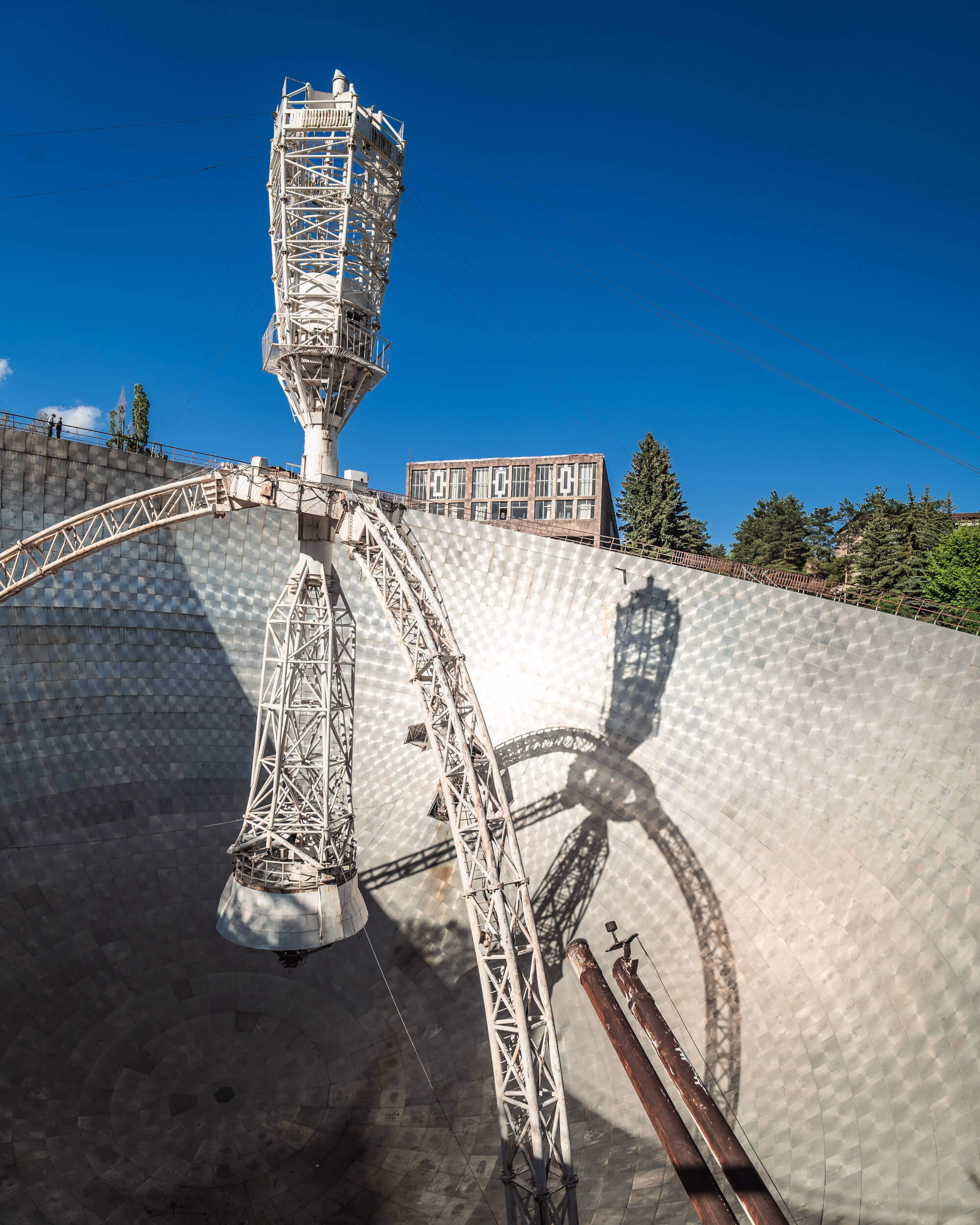 An abandoned satellite dish with a tall metallic structure and shadow cast on its surface, under a clear blue sky.