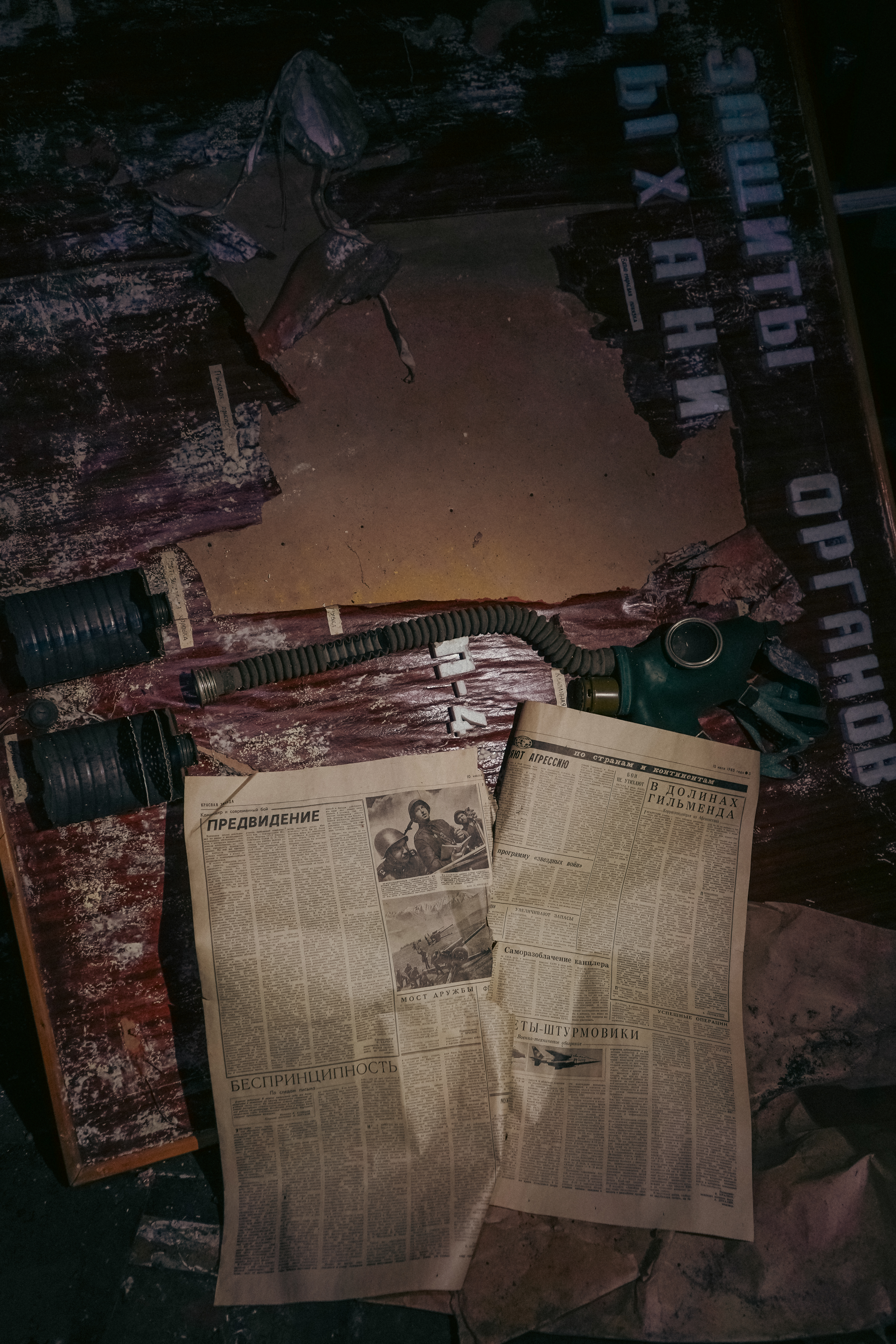 A green gas mask with tubing laid on a dark and worn wooden table, adjacent to two open, crumpled newspapers showing faded text, surrounded by dust and debris.
