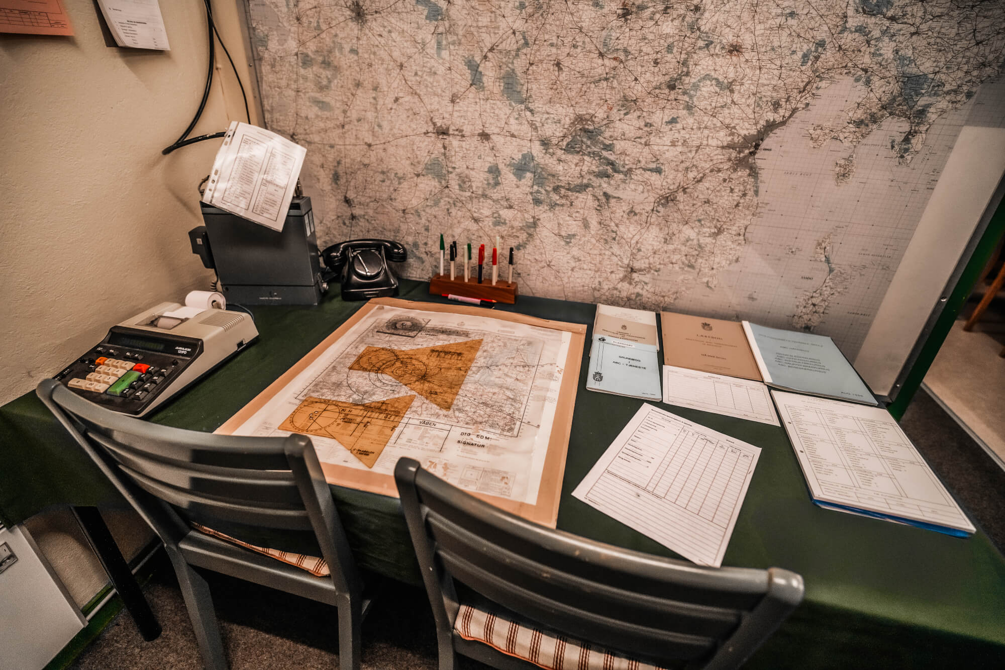 A desk with a vintage calculator, a classic black telephone, a large map on the surface, and various documents and colored markers organized on a table, set against a backdrop of maps on the wall.