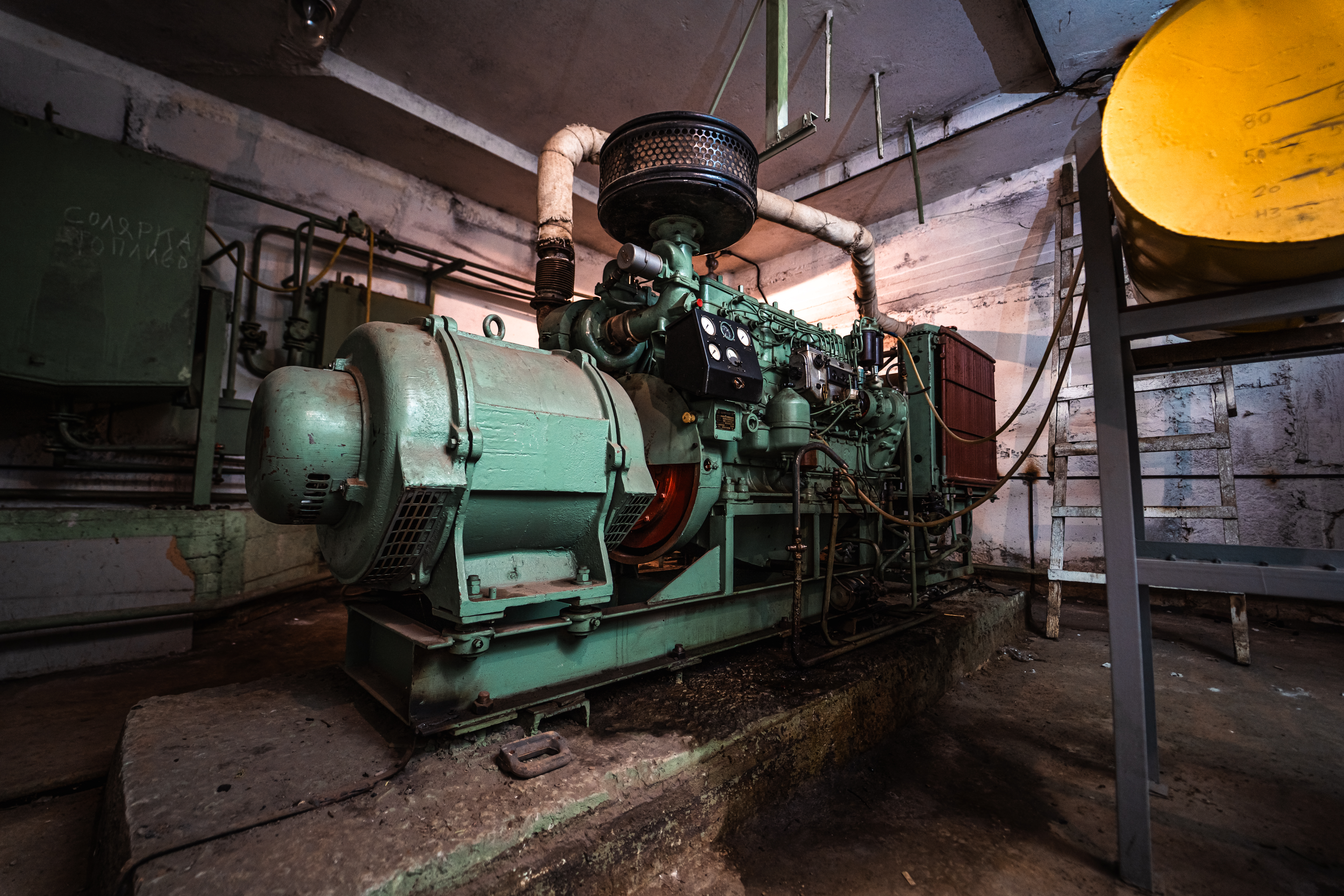 A green industrial generator situated in an unkempt room with visible pipes and an adjacent yellow tank. The scene is dimly lit, emphasizing the age and wear of the equipment.
