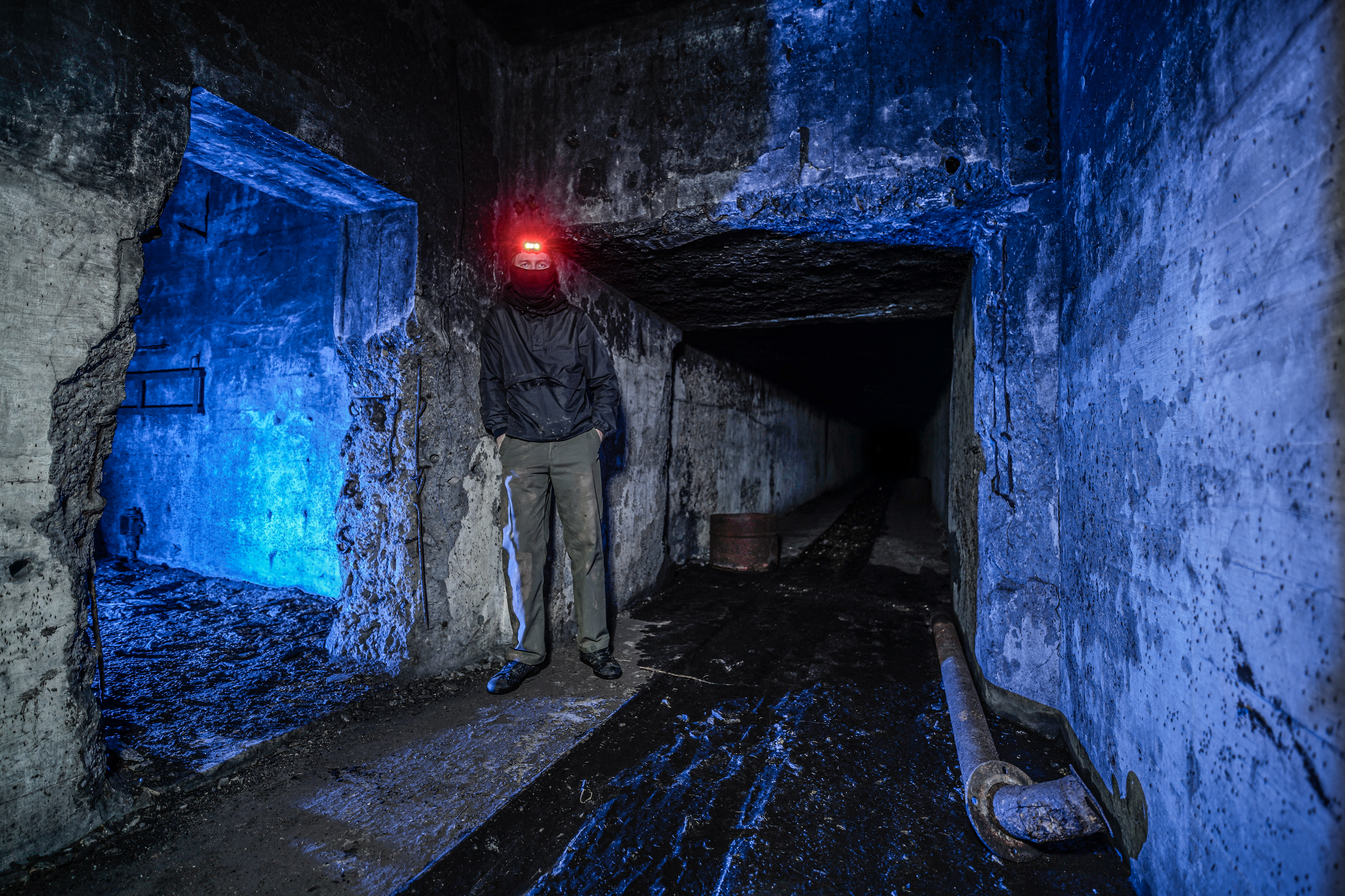 A person in dark clothing stands in an underground tunnel with a red headlamp, surrounded by concrete walls illuminated in blue light. A dark passageway extends into the background.