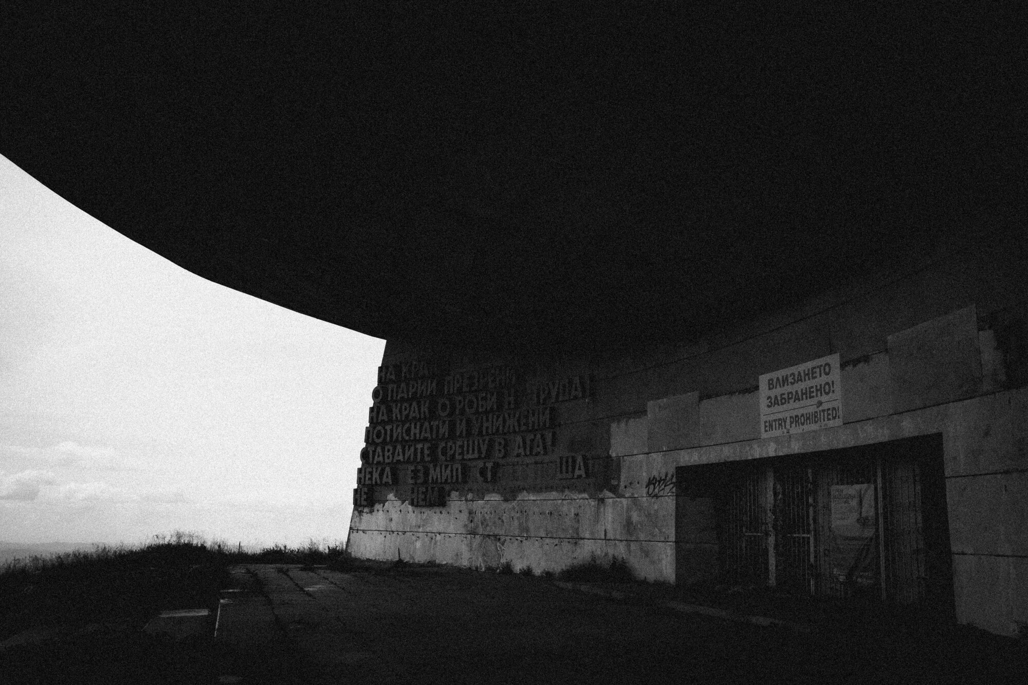 An abandoned building with a large overhanging roof, partially obscured in shadow. The walls feature faded inscriptions, and there is a sign stating 'Entry Prohibited.' The sky above is overcast, with a grassy area in the foreground.