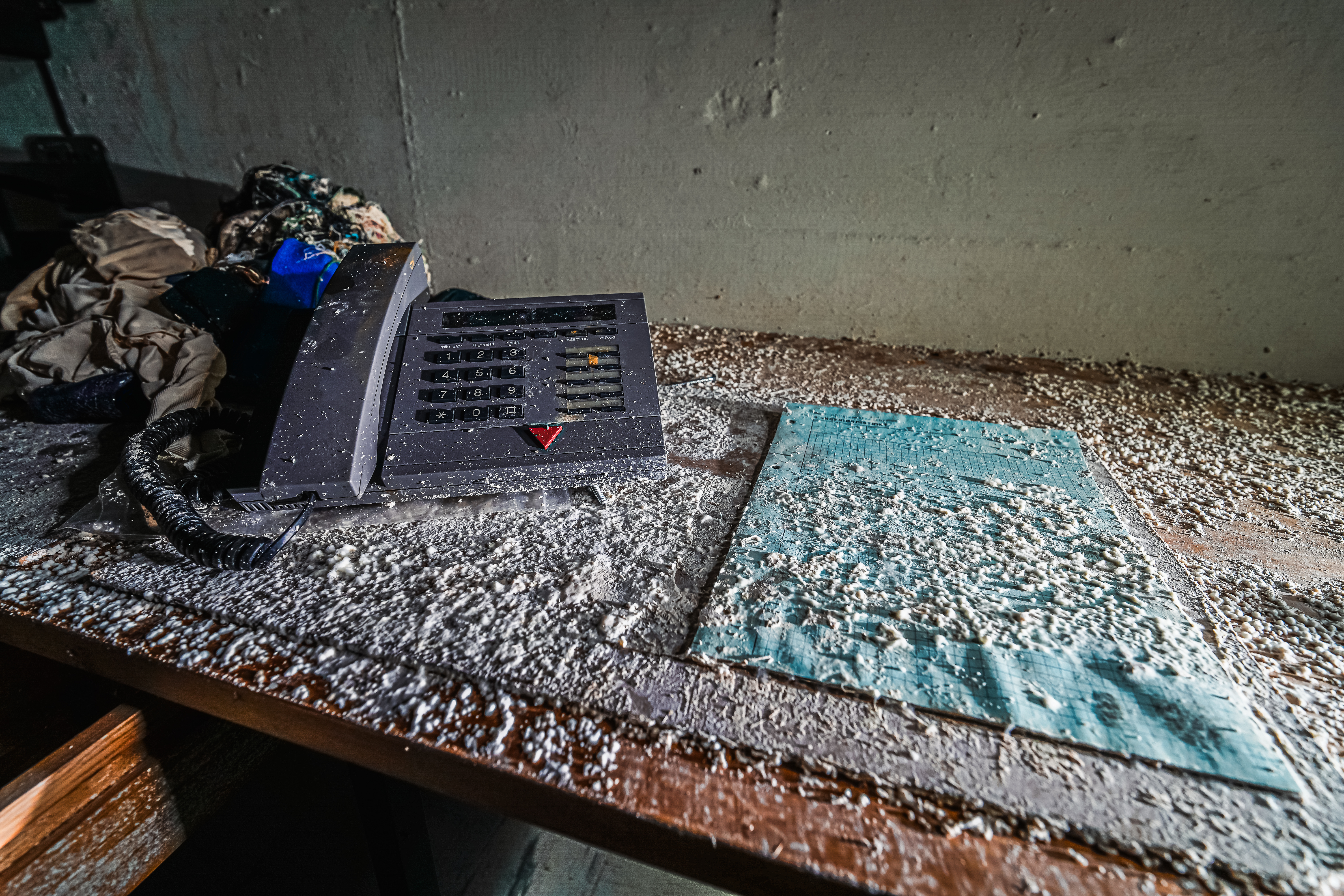 An abandoned office desk with an old telephone covered in dust and debris, accompanied by crumpled clothing and a piece of blue graph paper.