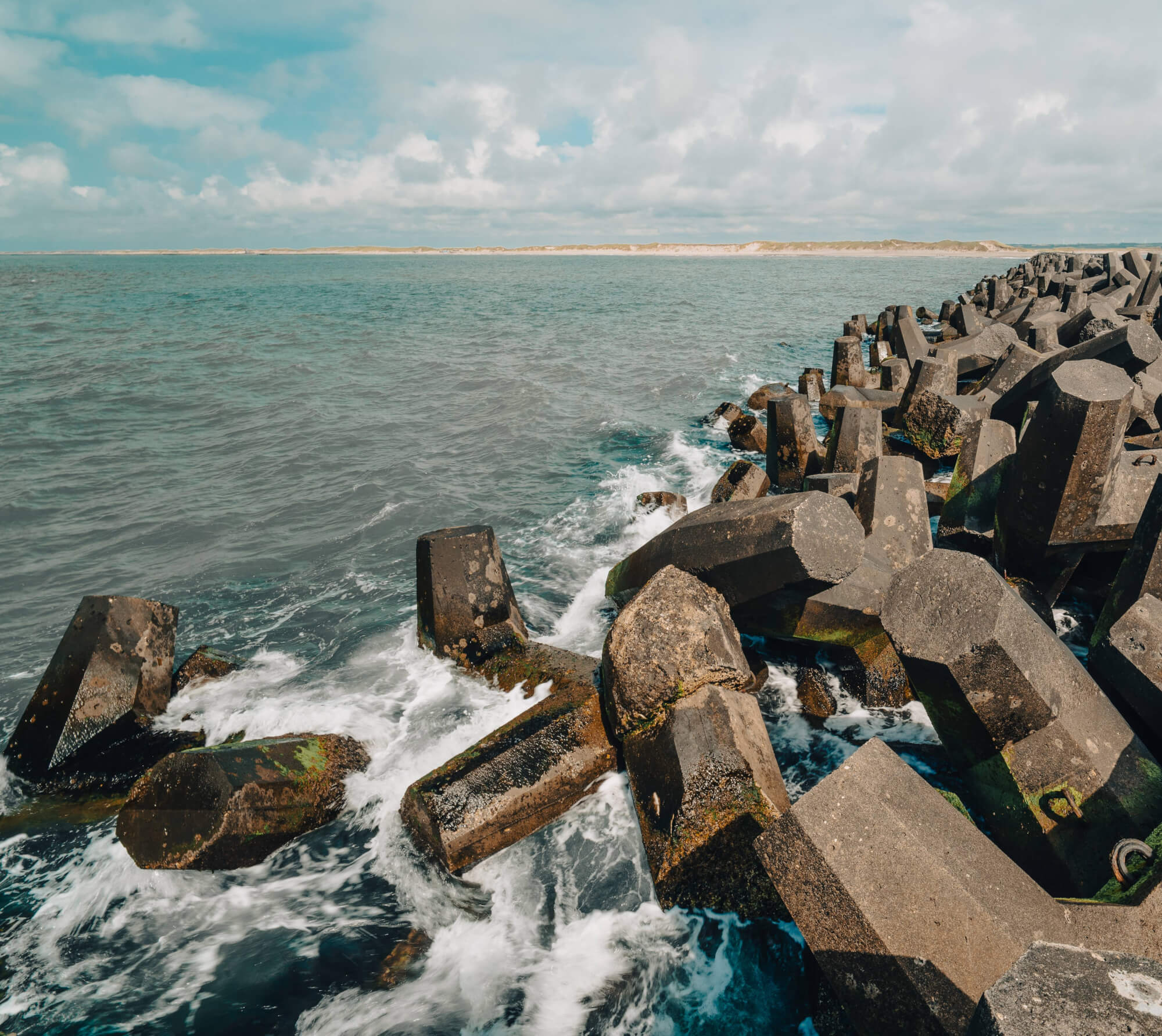 A coastal jetty with large concrete blocks partially submerged in water, surrounded by waves. The background features a blue sky with clouds and a distant sandy beach.