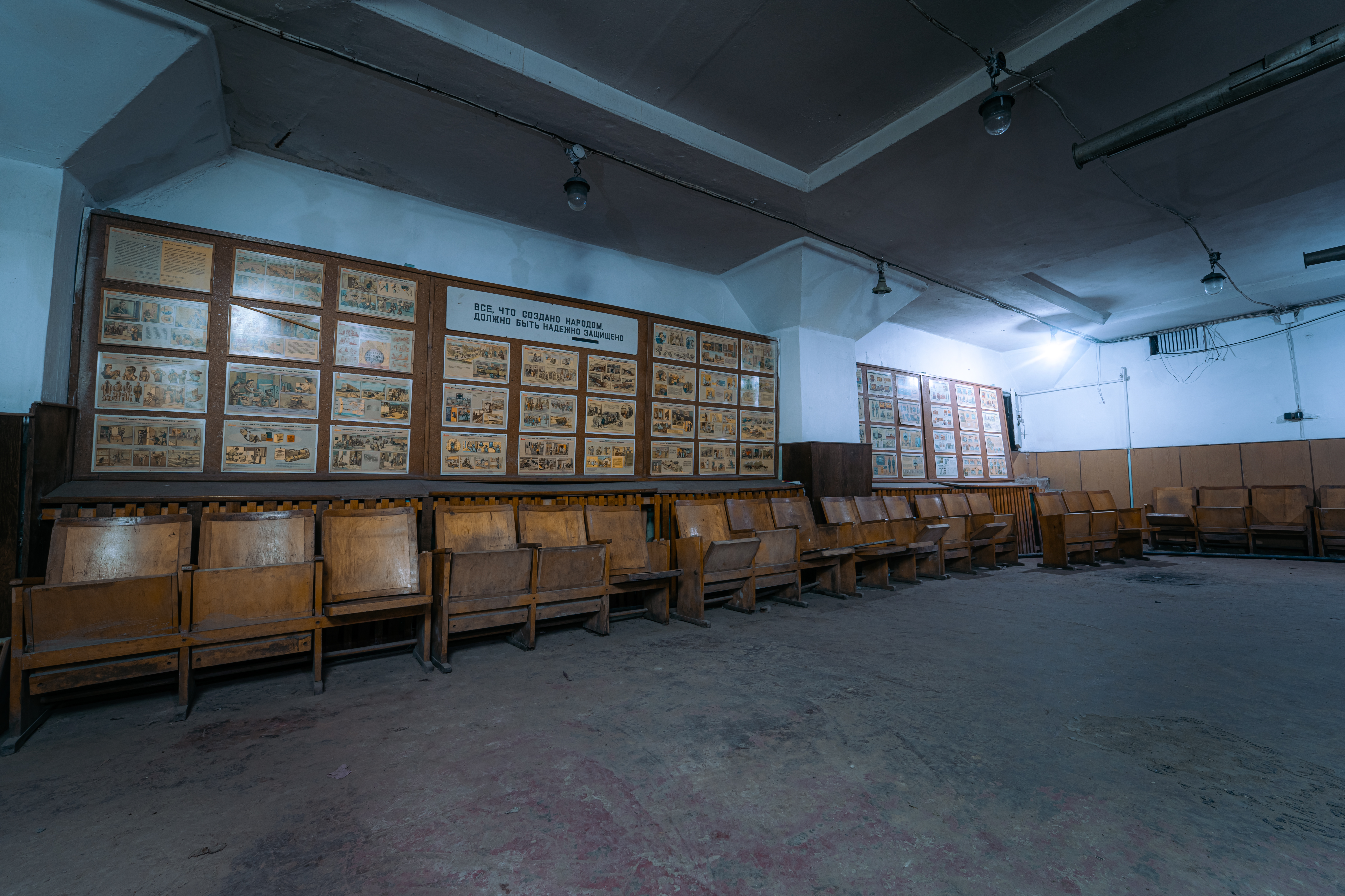 A deserted waiting room with wooden benches in rows and walls covered with illustrated panels and text, creating a somber and nostalgic ambiance.