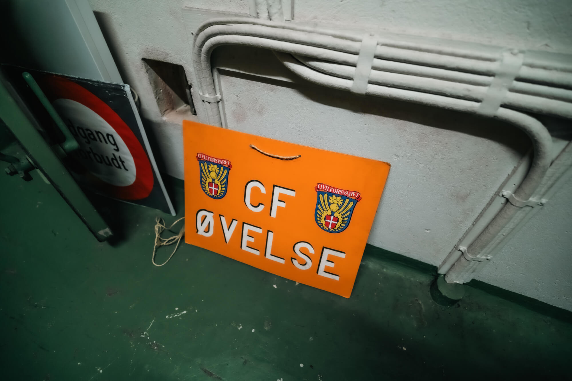 An orange sign with the text 'CF ØVELSE' rests on a green floor beside a round traffic sign and utility pipes against a concrete wall.