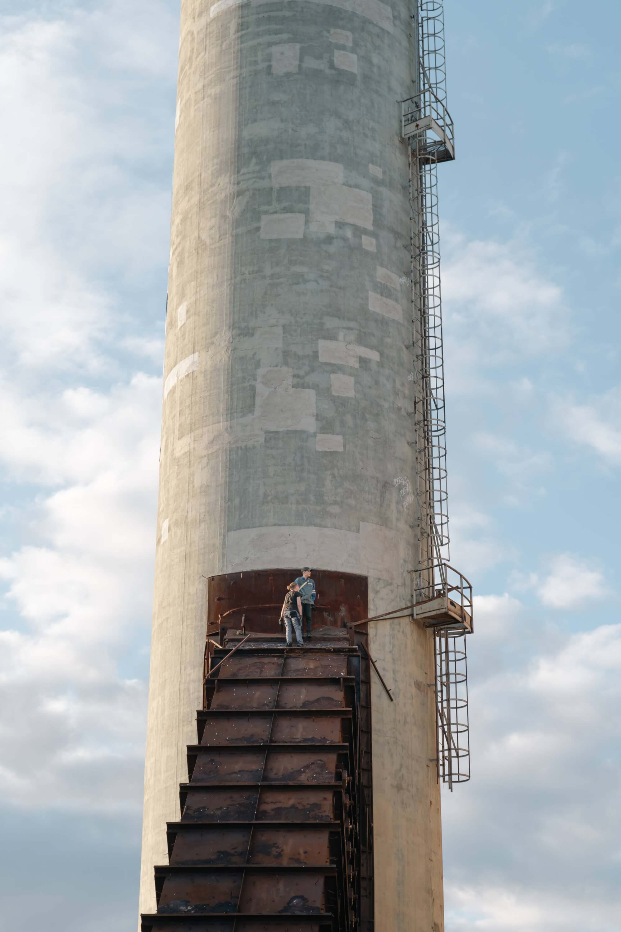 Two people standing on a rusted metal platform at the top of a large concrete smokestack, with a blue sky and clouds in the background.