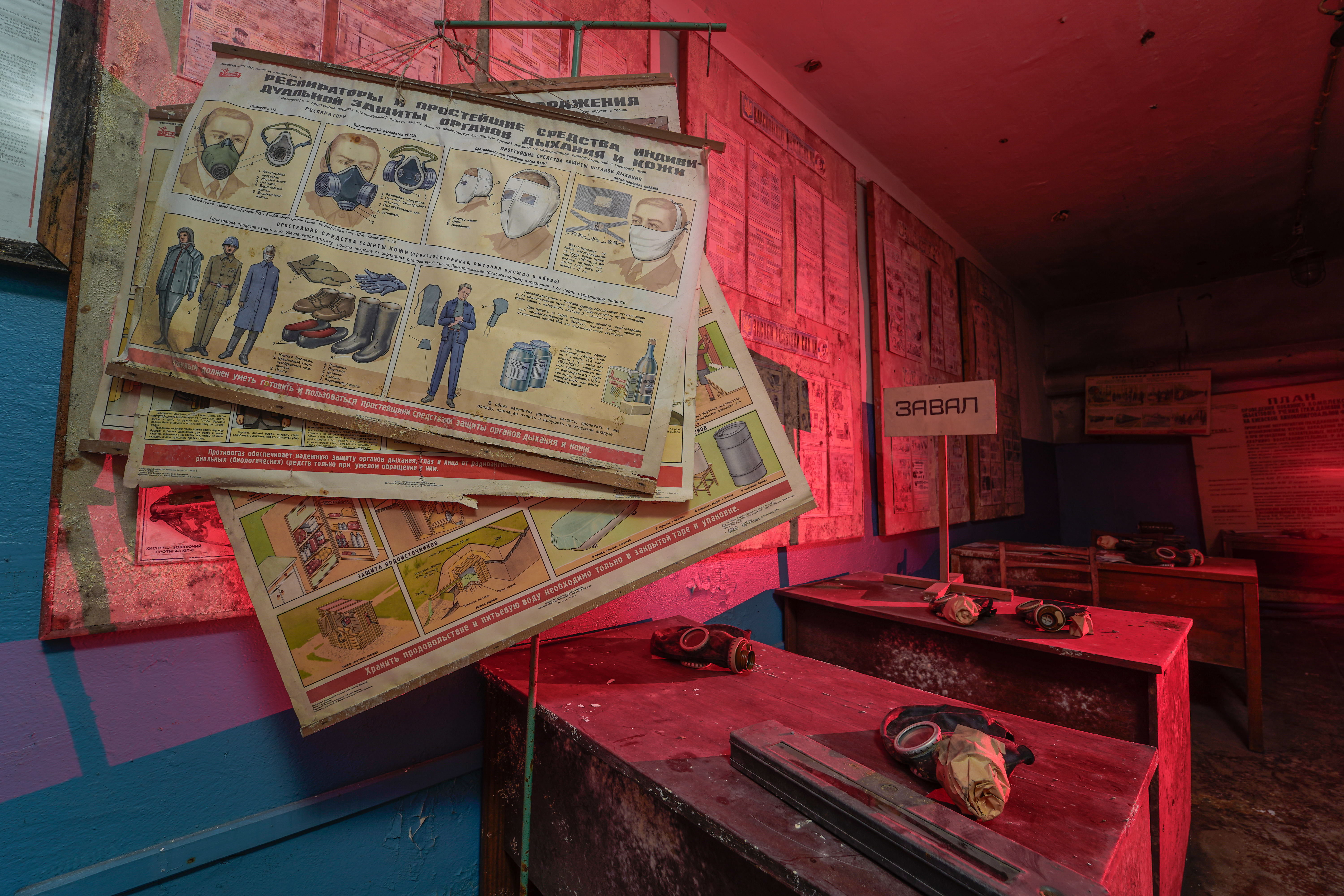 An interior space with red lighting featuring posters on the walls about protective equipment. Wooden tables hold gas masks and other safety gear, suggesting an abandoned emergency preparedness area.