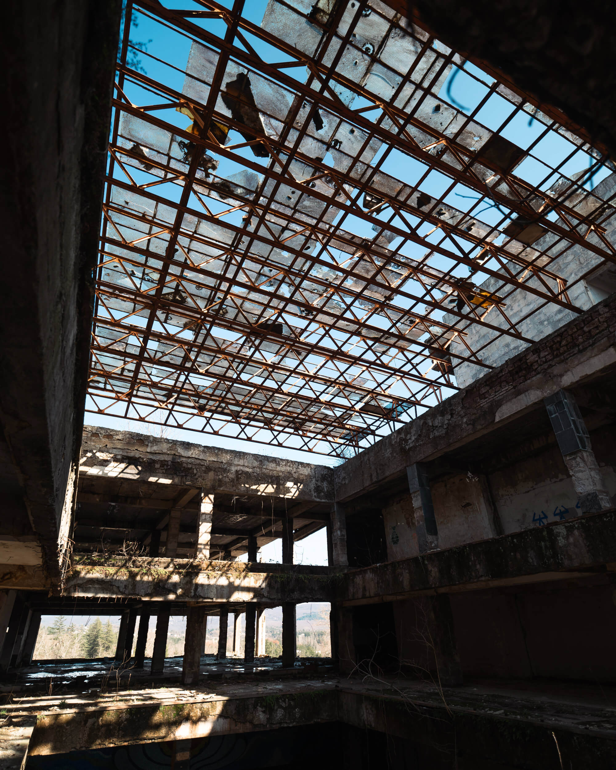 A dilapidated interior of an abandoned building with a rusty roof and fragmented skylights. Sunlight filters through the openings, revealing overgrown vegetation and shadowy corners in the concrete structure.
