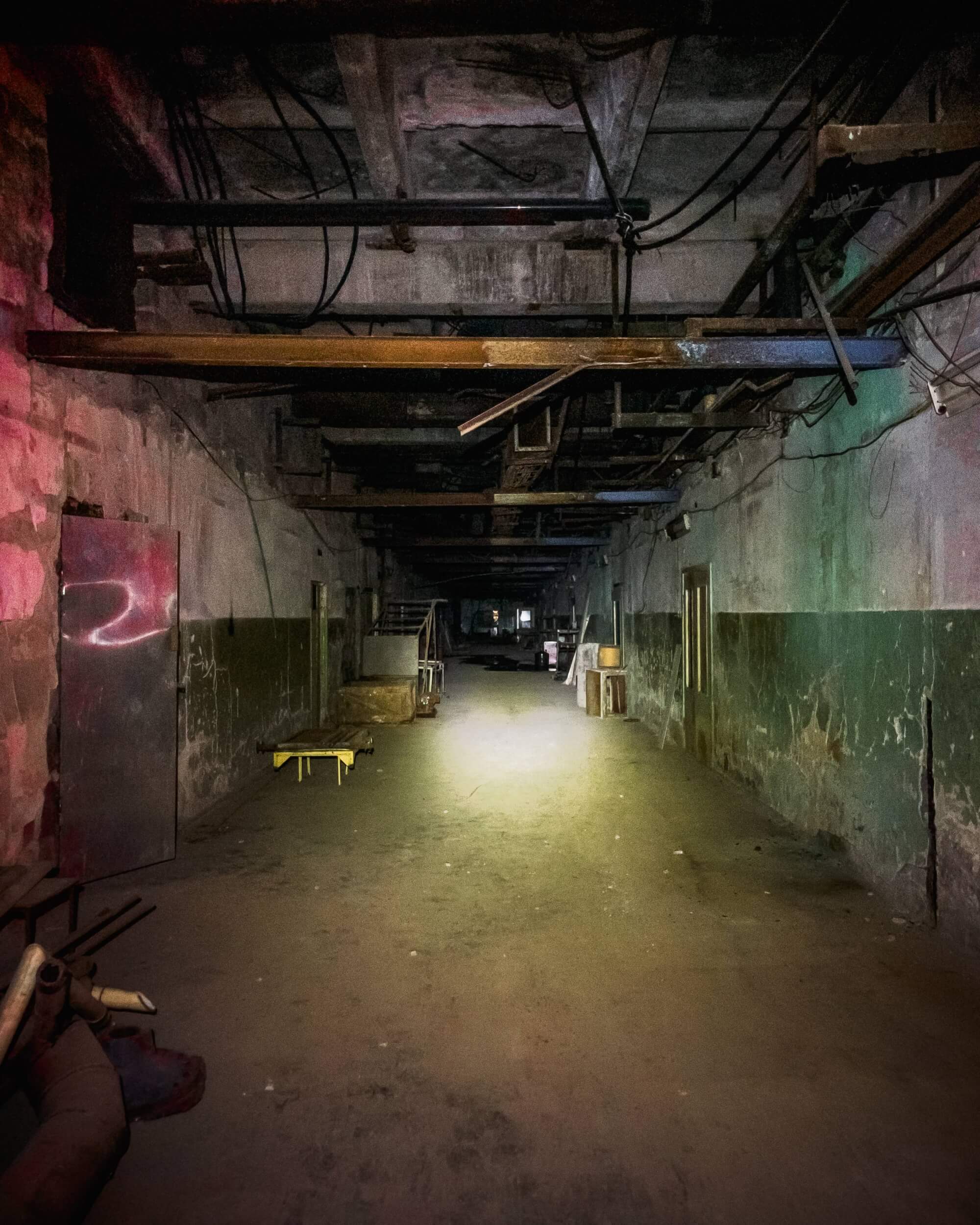 A long, empty hallway with dim lighting, featuring dusty floors and scattered objects. The walls are partially visible in green and gray, and various shadows and debris create a haunting atmosphere.