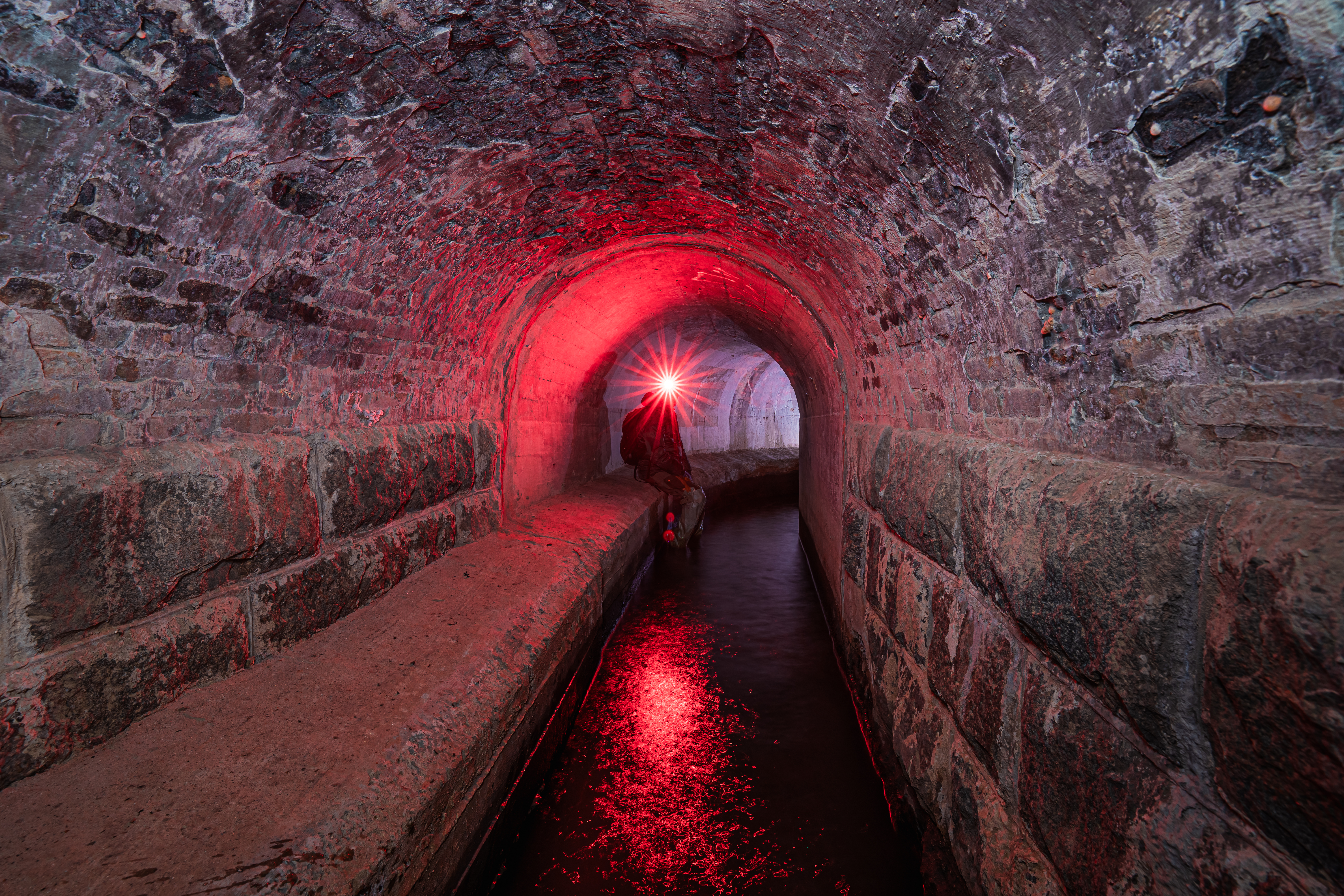A dimly lit underground tunnel with red lighting, featuring a figure sitting on a stone edge holding a flashlight, with water reflecting the light in the foreground.