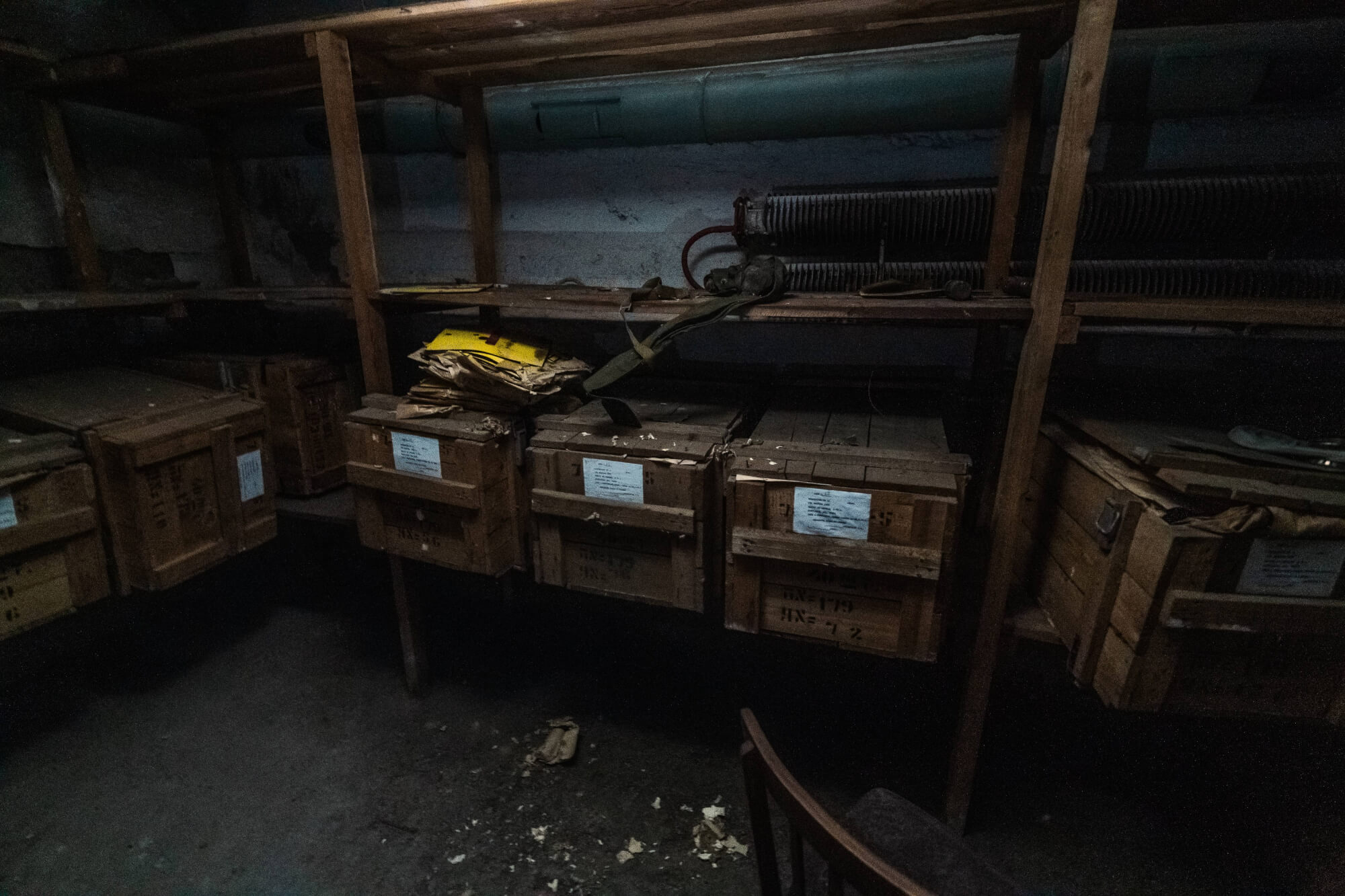 A dark storage room with wooden crates stacked on shelves, some containers lie open with papers spilling out. A single chair sits in the foreground, while the walls and shelves are dimly lit, creating a moody atmosphere.