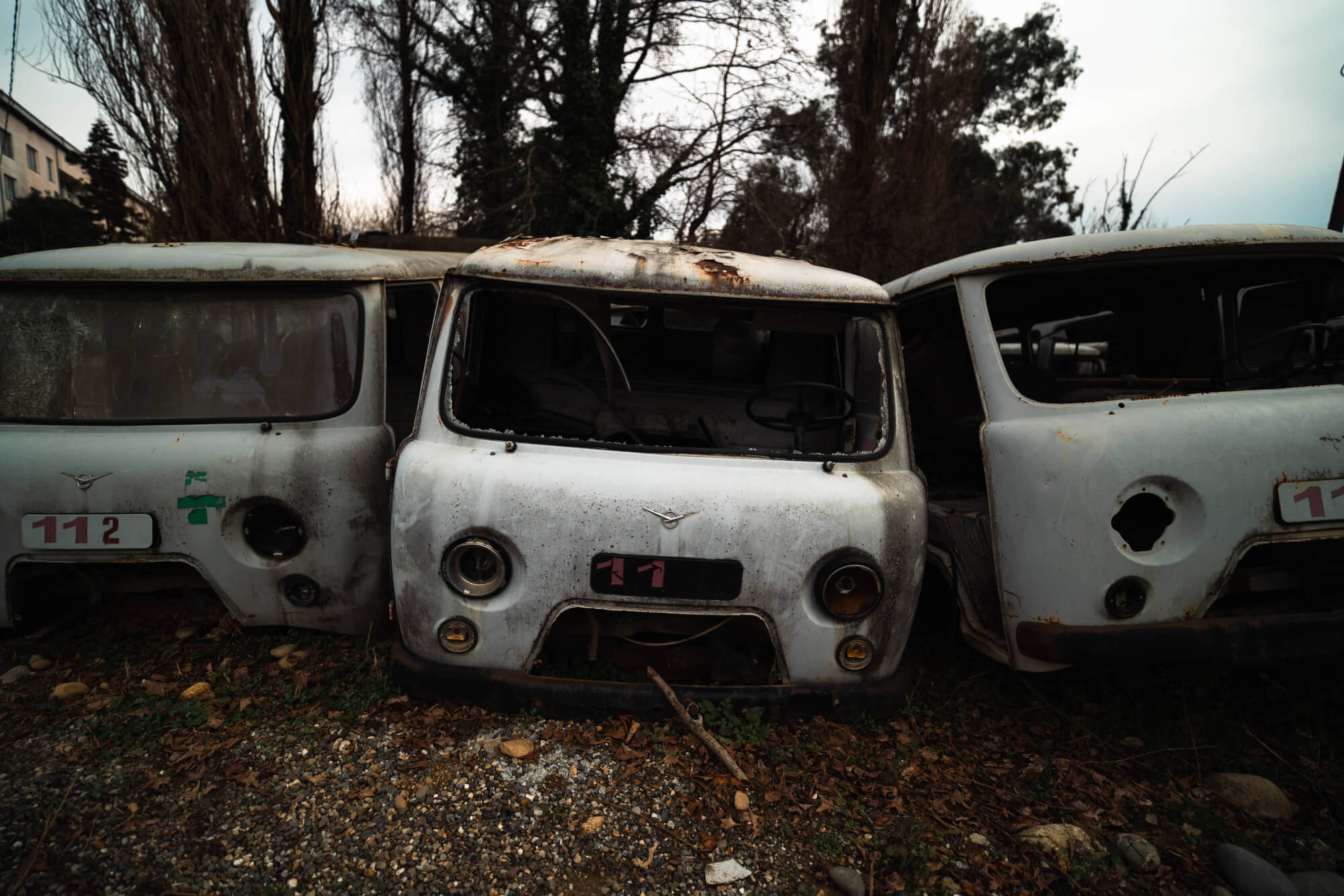 Three abandoned vintage vehicle fronts, displaying signs of rust and decay, set against an overcast sky and sparse vegetation.