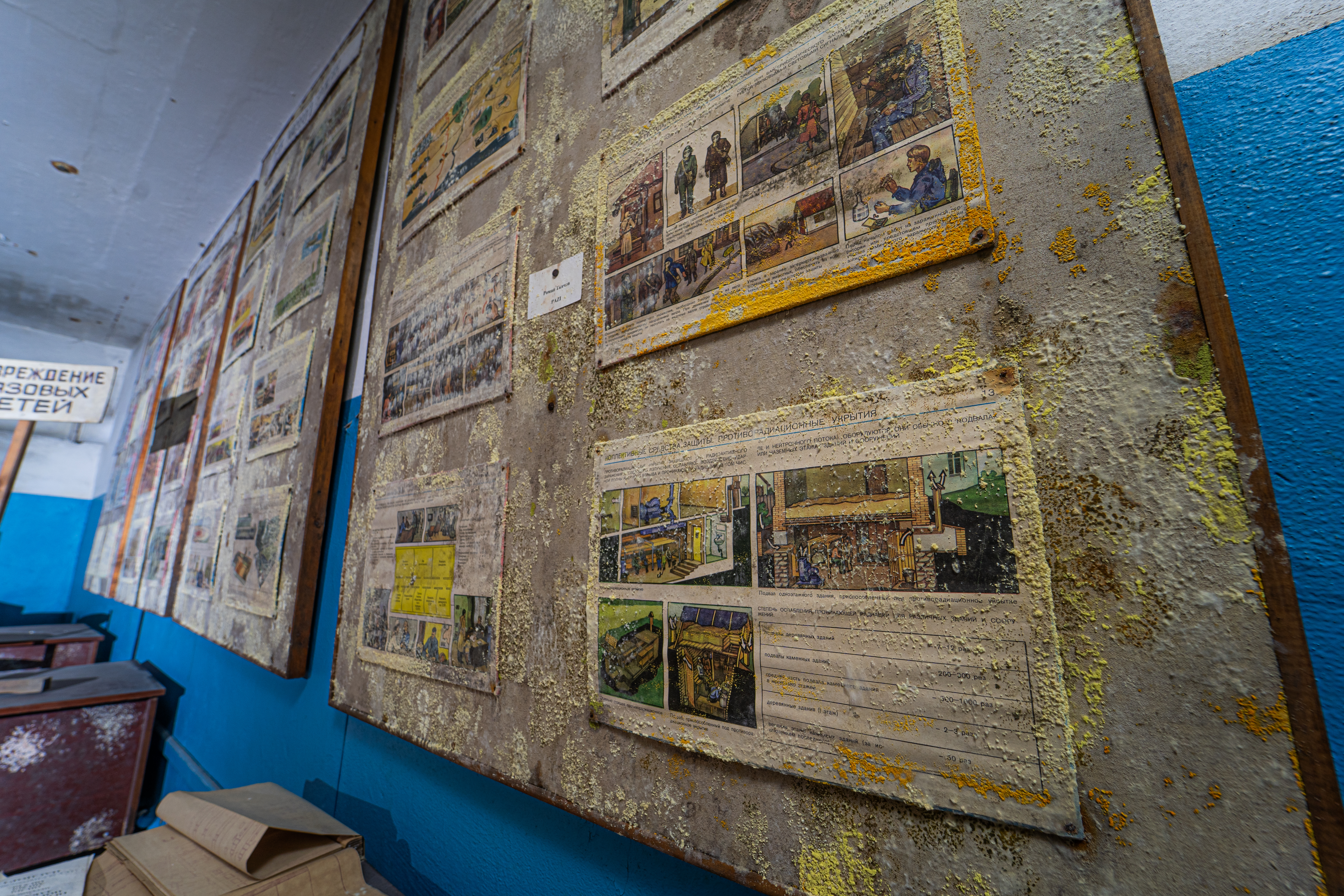 A close-up view of a wall covered with old, weathered posters and notices, with some areas showing growth. In the foreground, an open book rests on a dusty table.