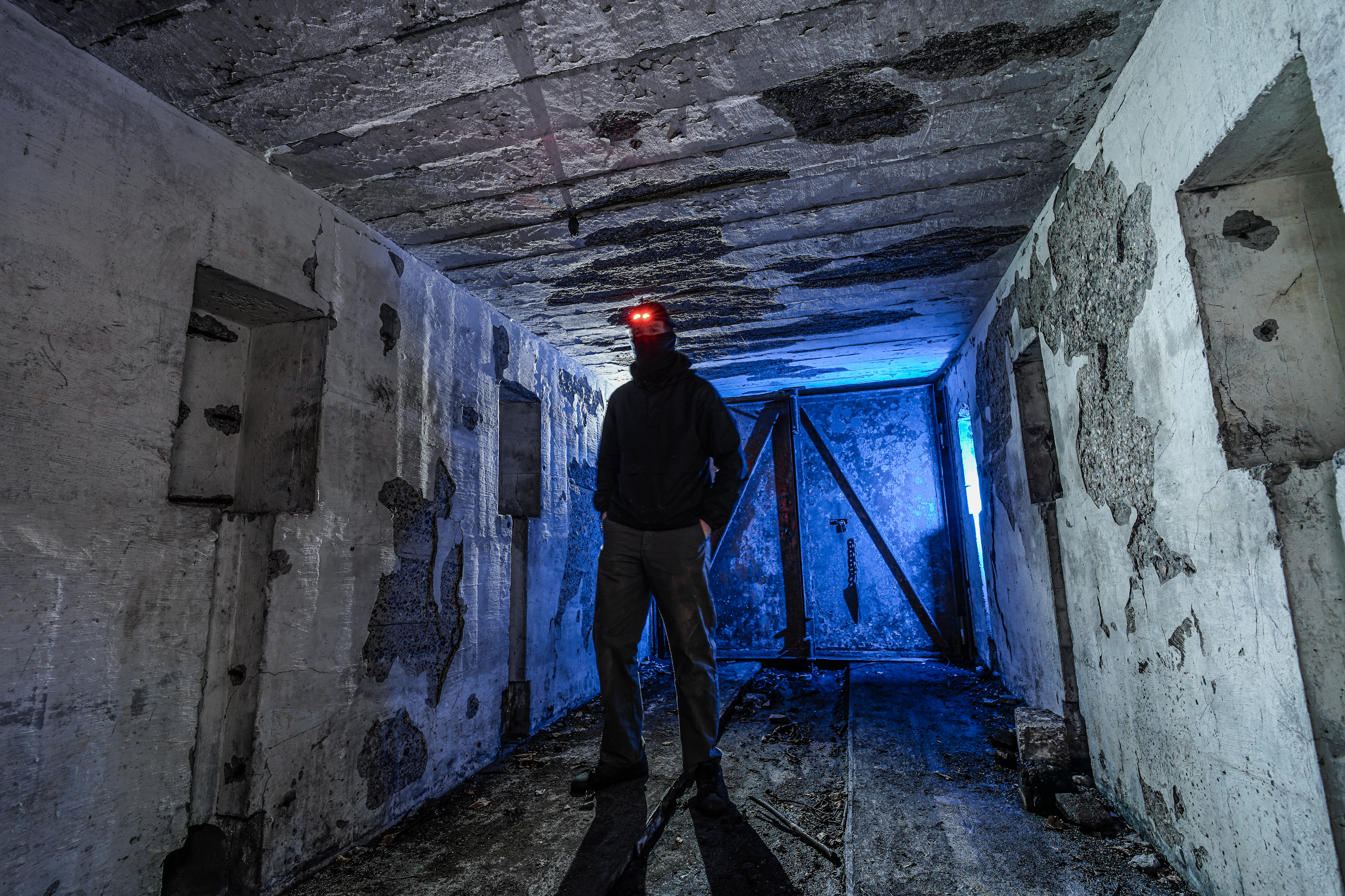 A person in a dark hoodie stands in an empty, dimly lit room with cracked concrete walls. The figure's headlamp glows red, contrasting with the blue light from a window. The floor is dirty and uneven, and there are small openings in the walls.