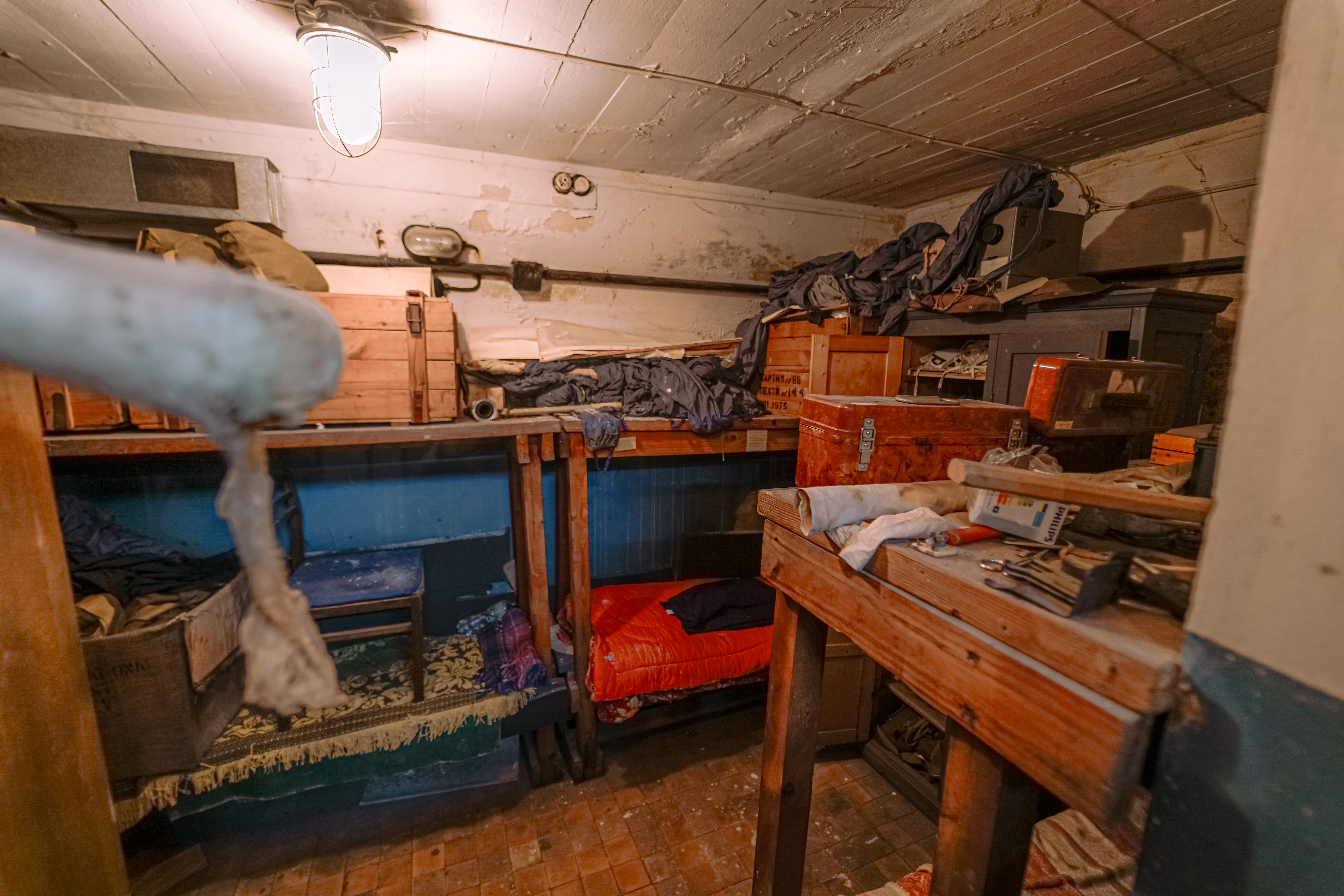 A cluttered basement with wooden shelves filled with boxes and old clothing. A red bed is visible against the wall, with a variety of objects and tools scattered on a nearby wooden table.