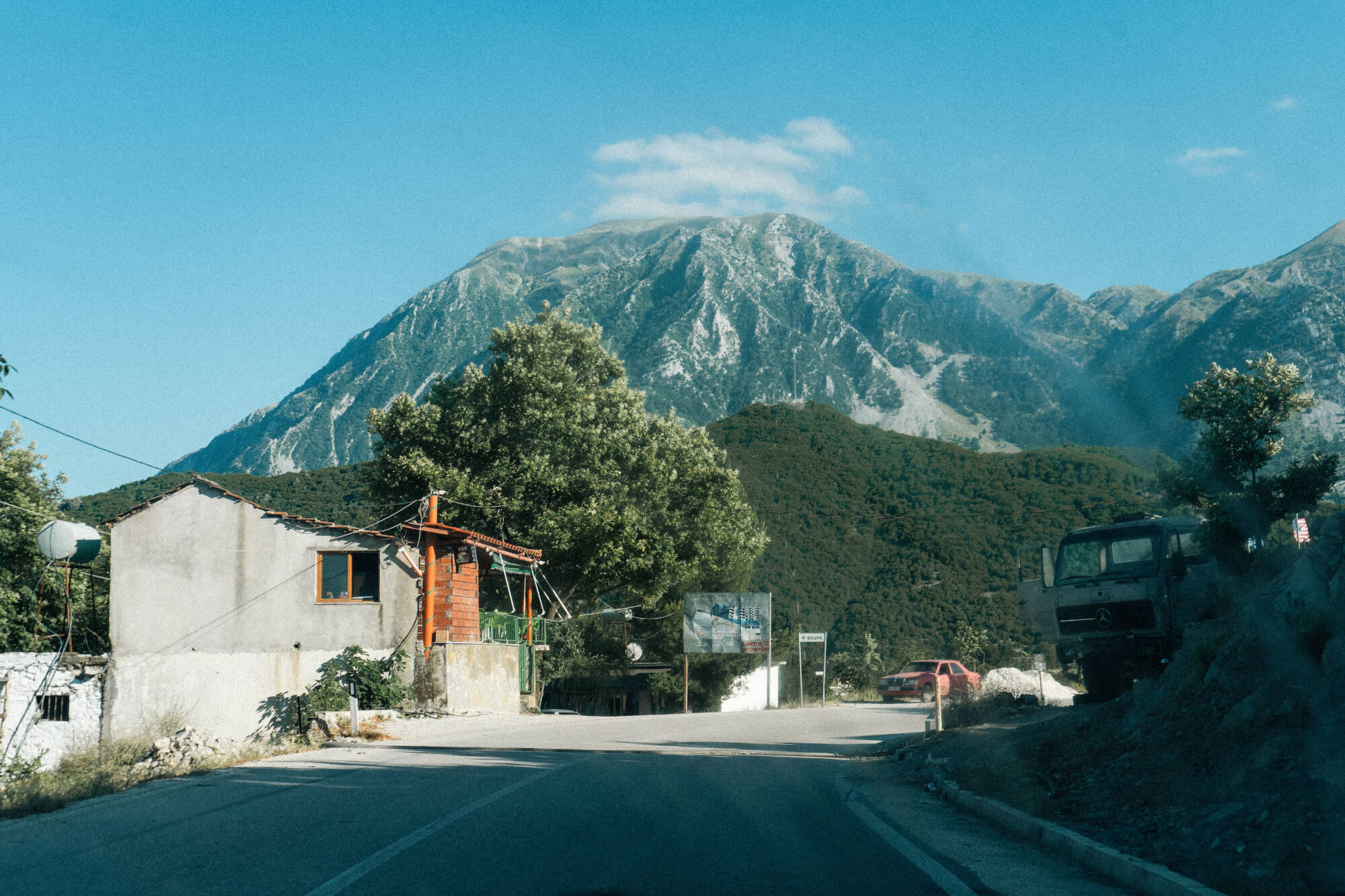 A rural house beside a road with mountains in the background. A tree is in front of the house, and an old truck is parked to the right. The sky is clear and blue.