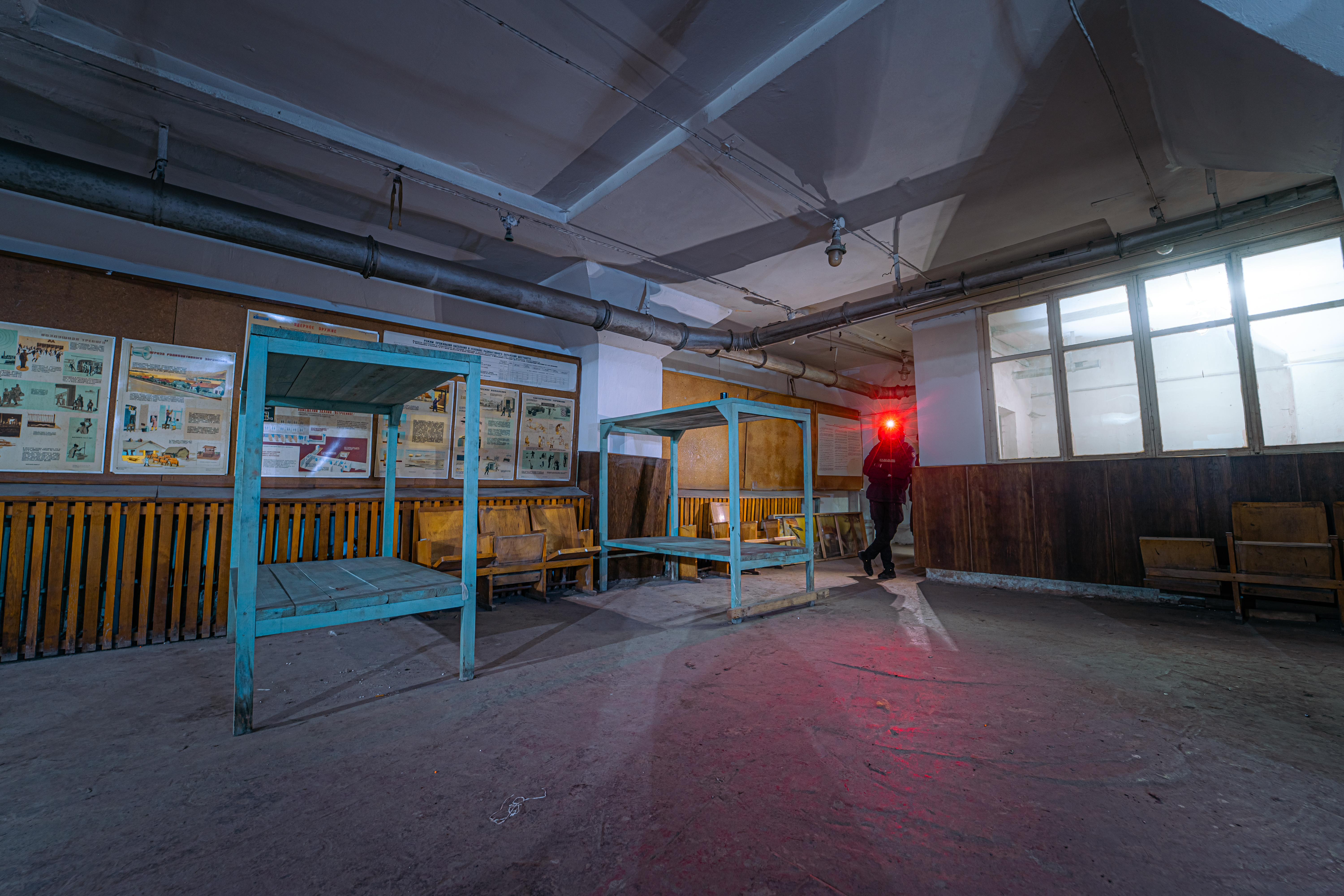 An abandoned room with empty teal frames and wooden benches, illuminated by the red light of a person carrying a flashlight. Vintage posters are visible on the walls in a dimly lit environment.