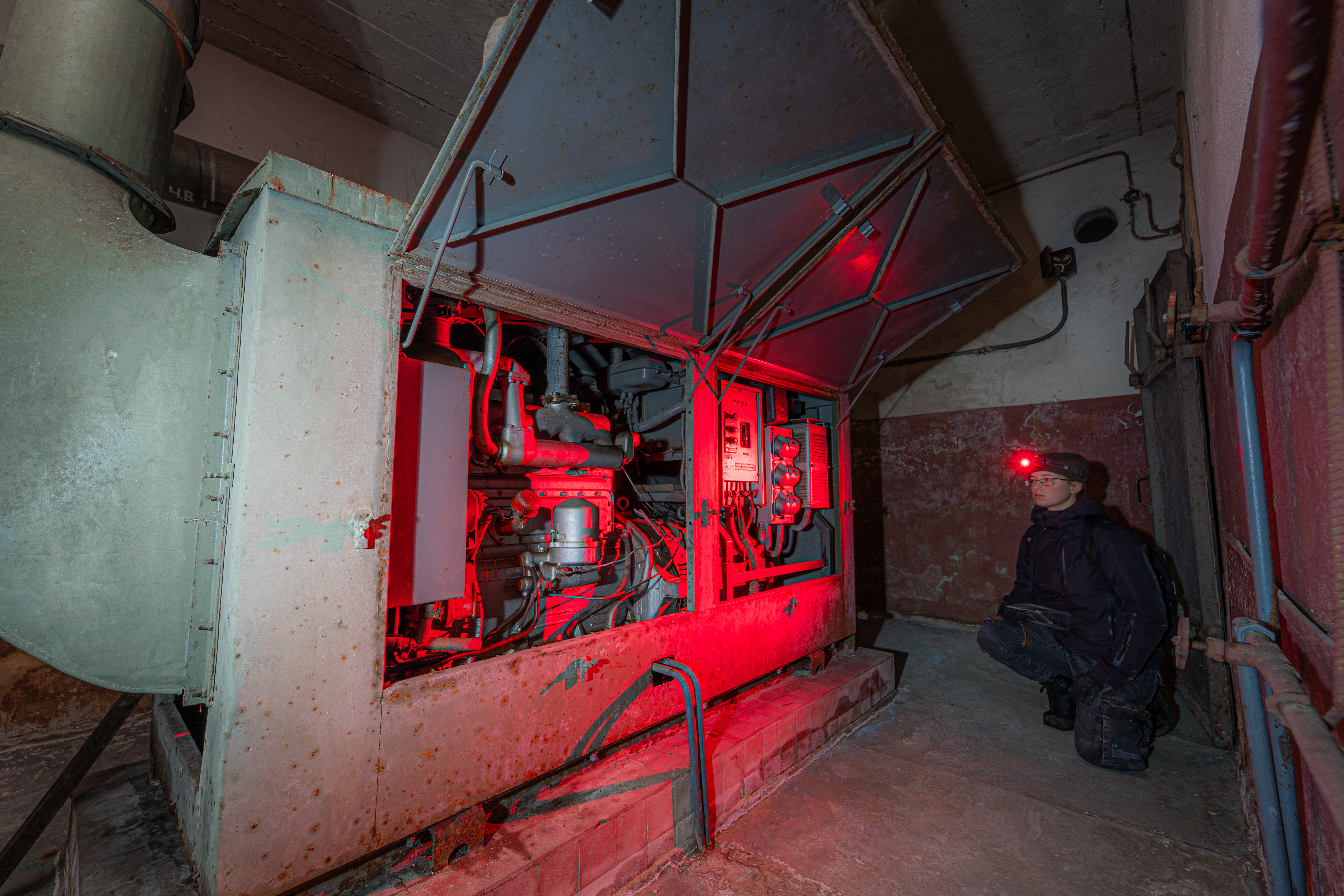 A person kneels next to an open generator in a dimly lit industrial space, illuminated by red lights, surrounded by rusty walls and pipes.