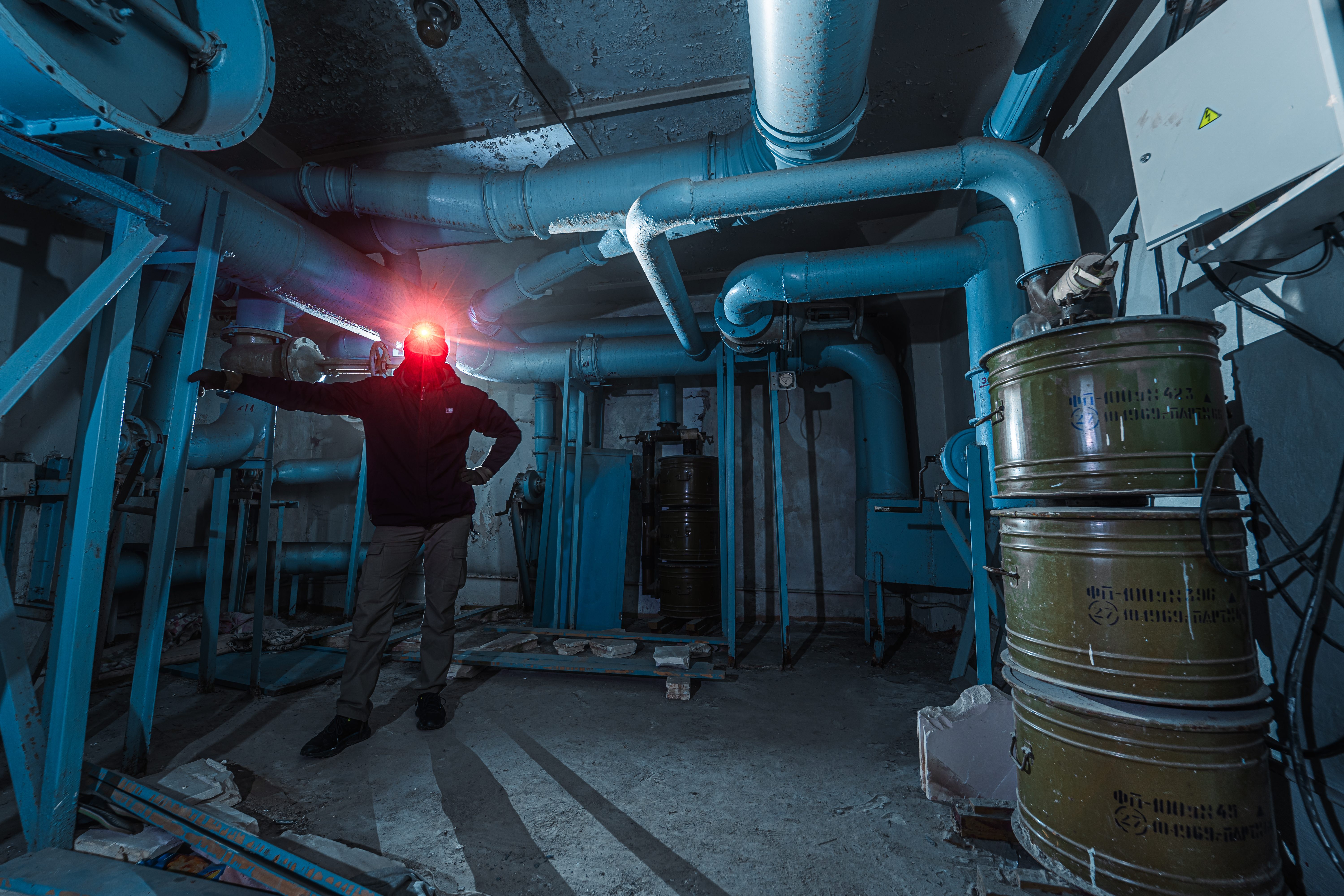 A person in a dark industrial room filled with blue pipes and rusty barrels, standing with one hand raised and illuminated by a headlamp.