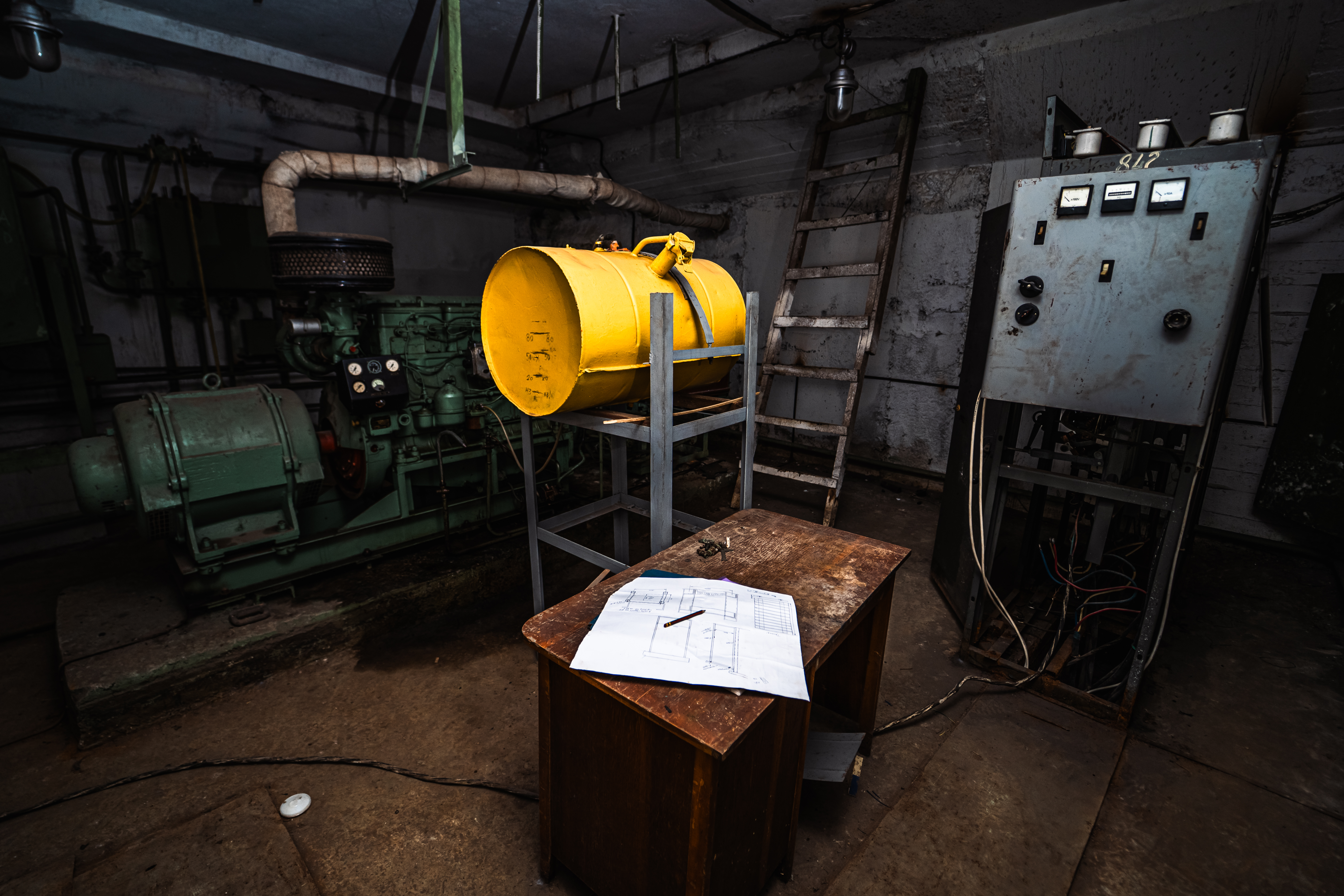 An industrial room featuring a yellow barrel on a metal stand, a wooden table with technical drawings, a rusted green machinery in the background, and a leaning wooden ladder.