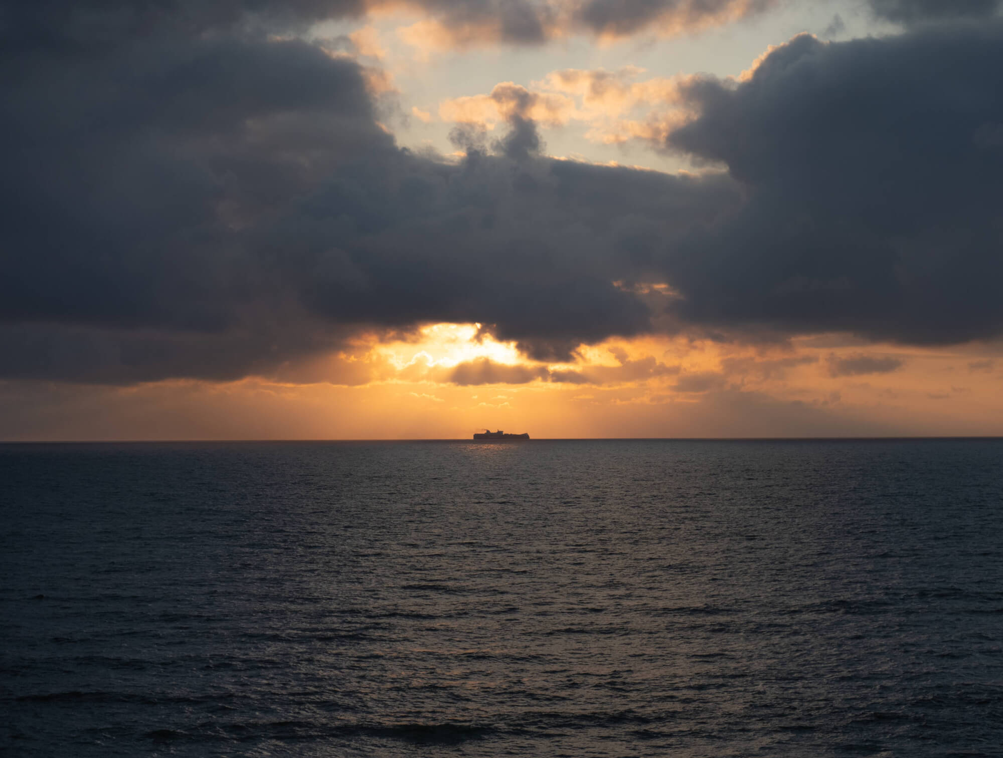 A distant cargo ship is seen on the horizon during sunset, surrounded by dark clouds and a warm, colorful sky, over a calm sea.
