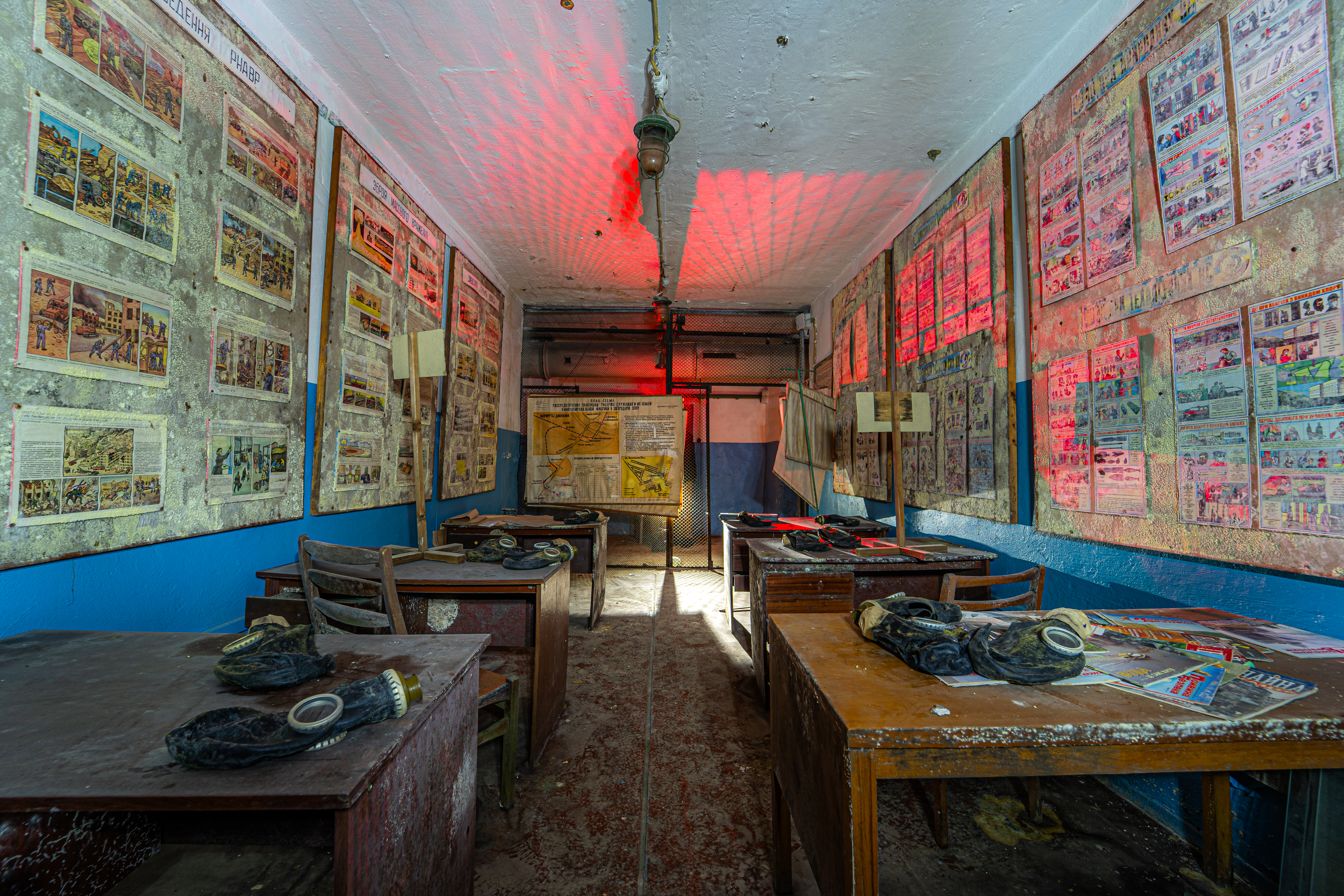 An abandoned classroom with wooden desks and gas masks on them. The walls are covered with faded educational posters, and there is a dim red light illuminating part of the room. Dust particles are visible in the air.