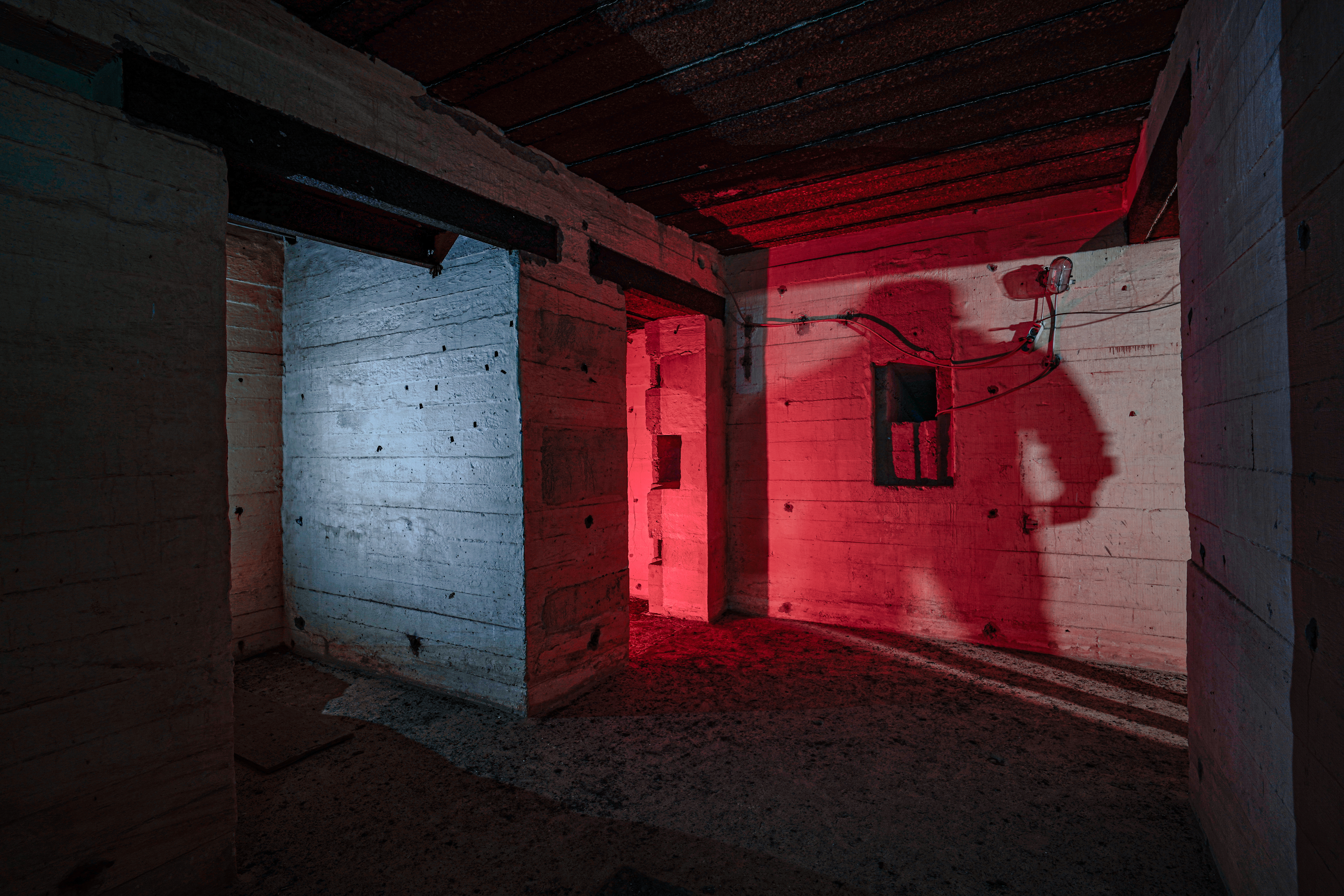 An abandoned room with concrete walls, illuminated by blue and red lights, casting long shadows. There are doorways and windows along the walls, enhancing the eerie atmosphere.