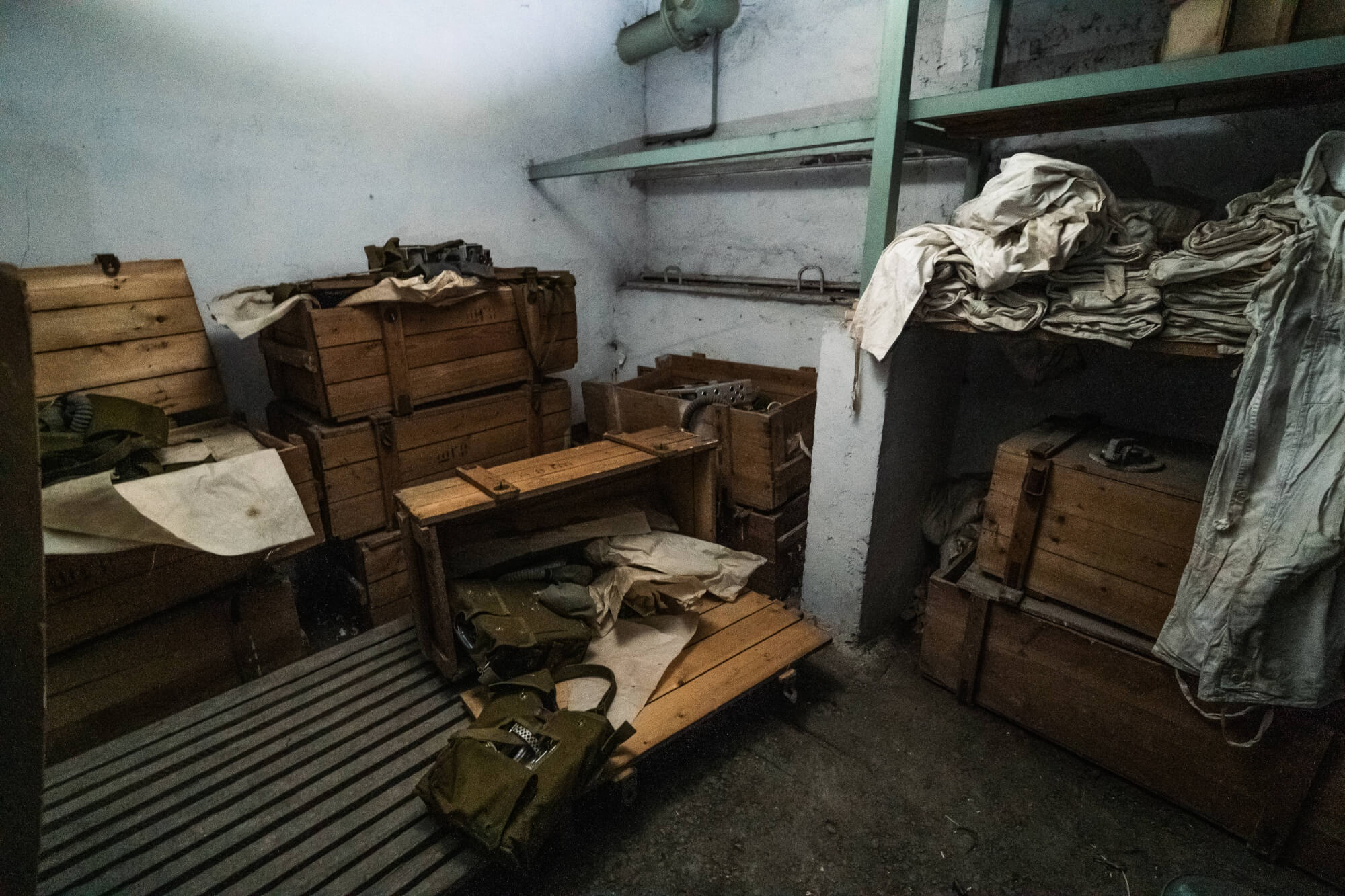 A dimly lit storage room filled with wooden crates and military gear. The floor is wooden slats, and there are white fabrics draped over some items. The walls are bare and peeling.