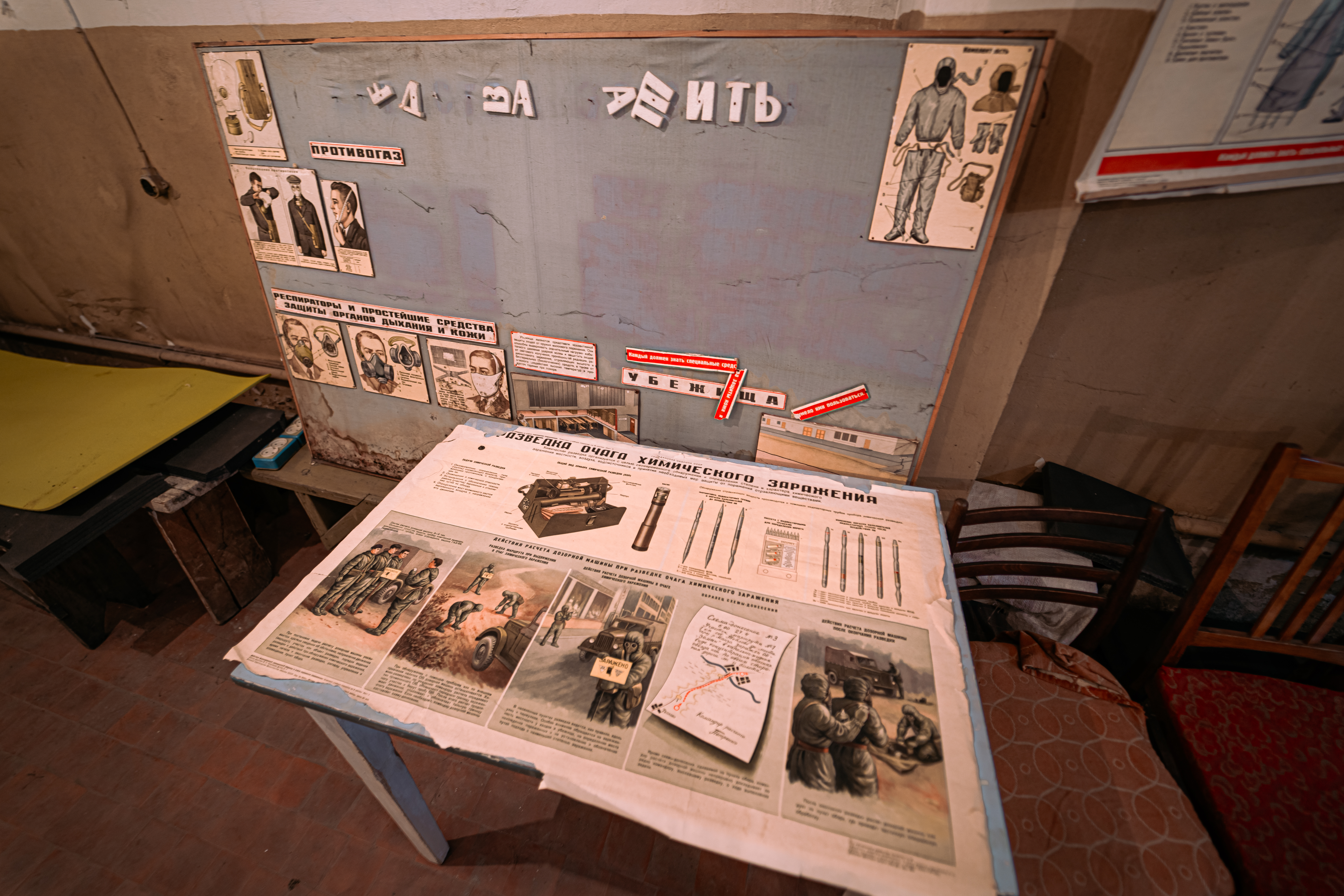 A display board with safety instructions and illustrations about gas masks and protective gear, alongside a table covered with informational sheets about chemical exposure. Chairs are scattered around, with a backdrop of a muted, aging wall.