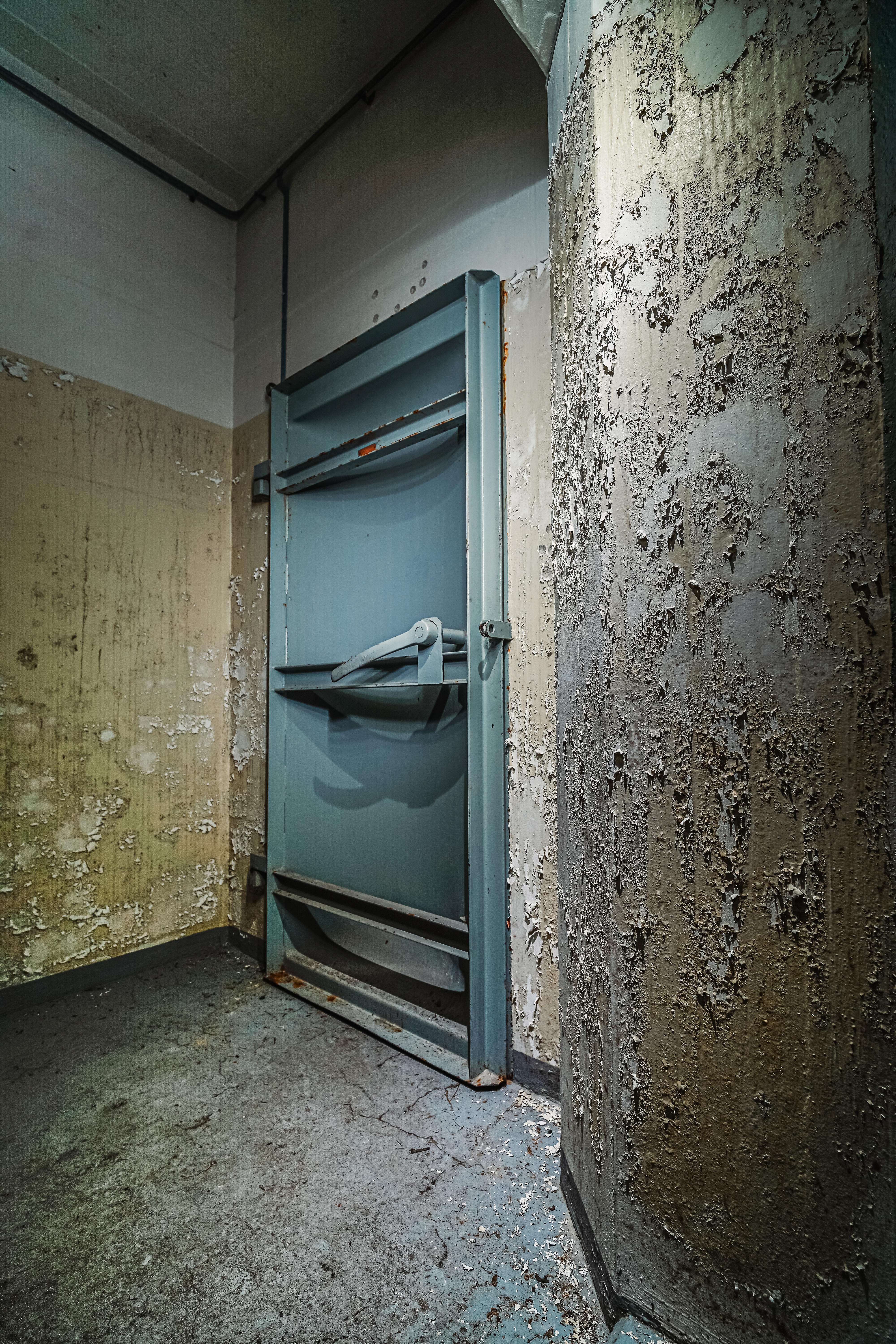 A partially open rusted metal door in a corner of a room with peeling paint and damaged walls, a scattered debris on the floor.